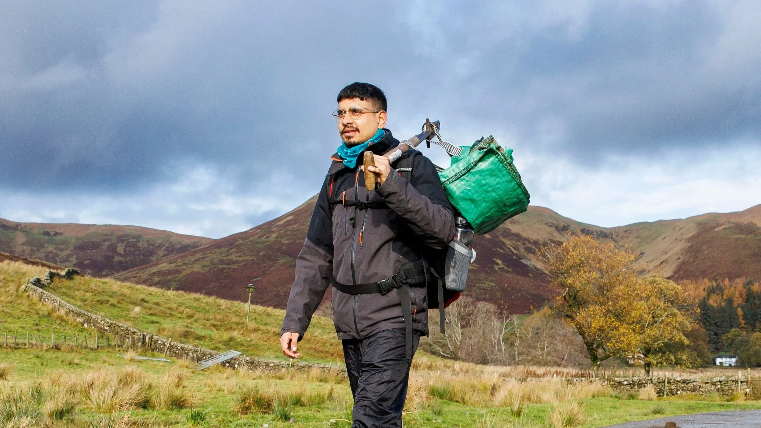 A man hiking on a muddy trail in a rural landscape, carrying a backpack and a green bag, with hills and trees in the background under a cloudy sky.