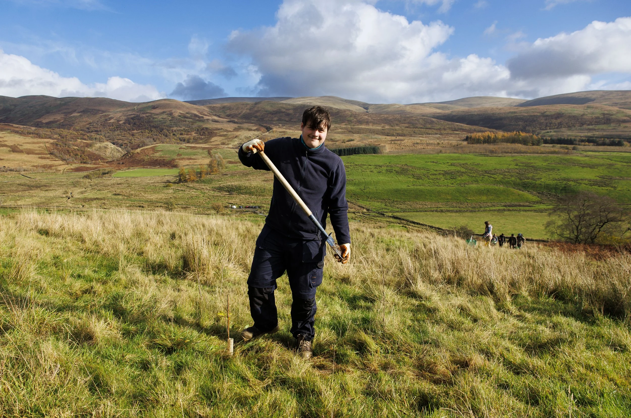 Group of people hiking through grassy field, led by a woman with a backpack and walking stick, surrounded by autumn foliage.