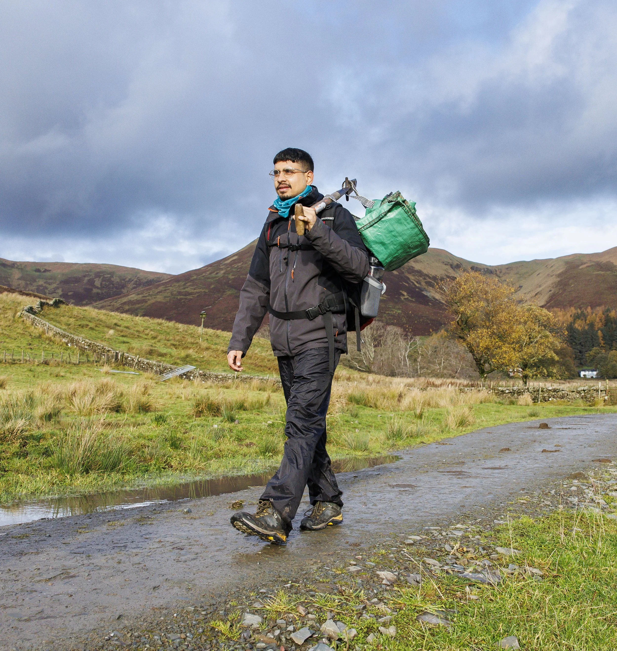 A young man walking towards the camera in the countryside with a rucksack on and carrying tree planting equipment. 