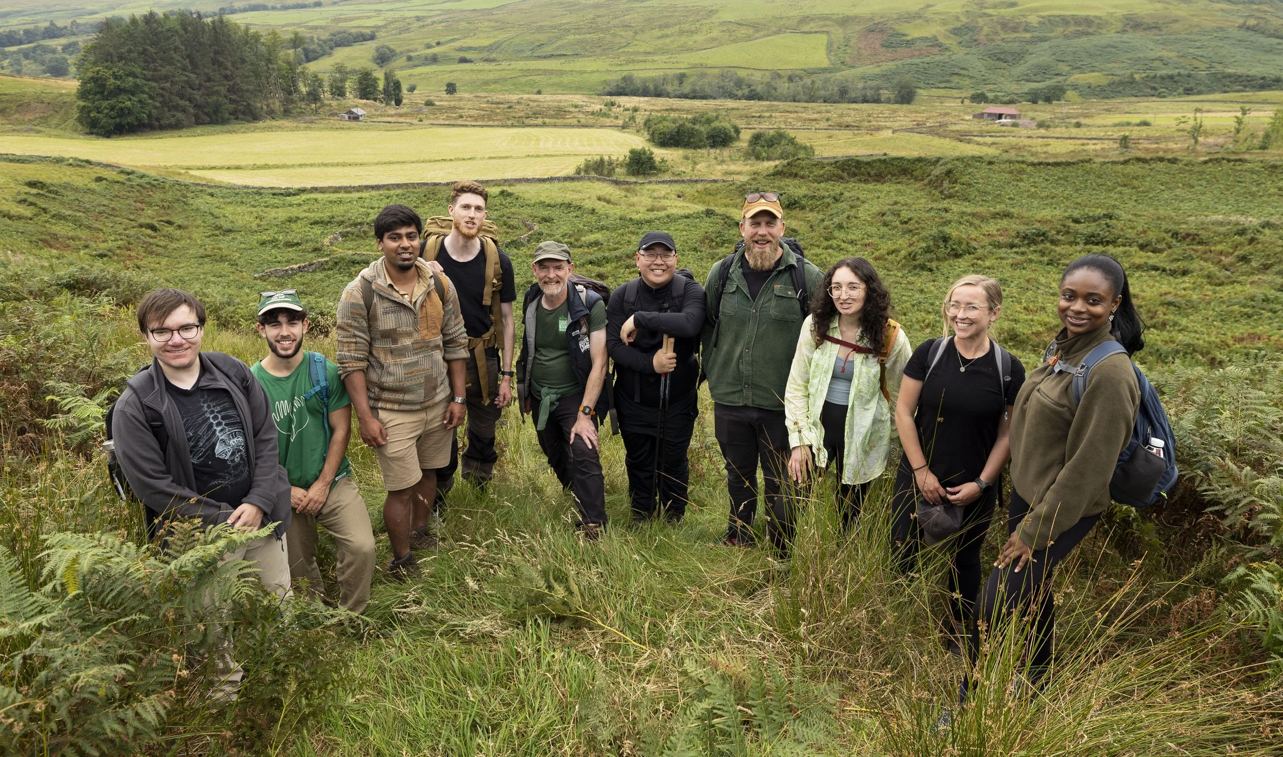 Group of ten diverse hikers standing outdoors in a lush, green landscape with rolling hills and fields in the background.