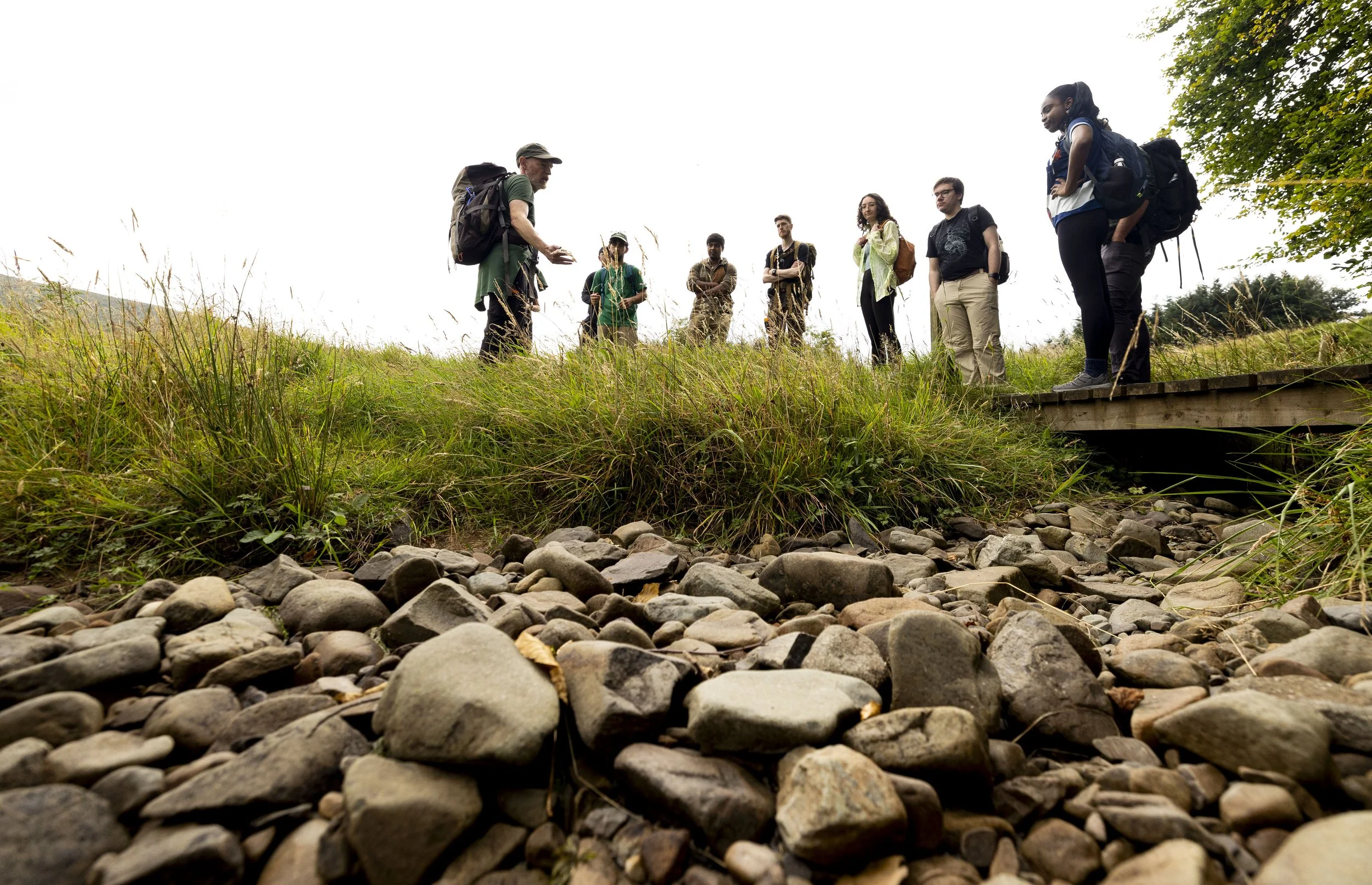 A man hiking on a muddy trail in a rural landscape, carrying a backpack and a green bag, with hills and trees in the background under a cloudy sky.