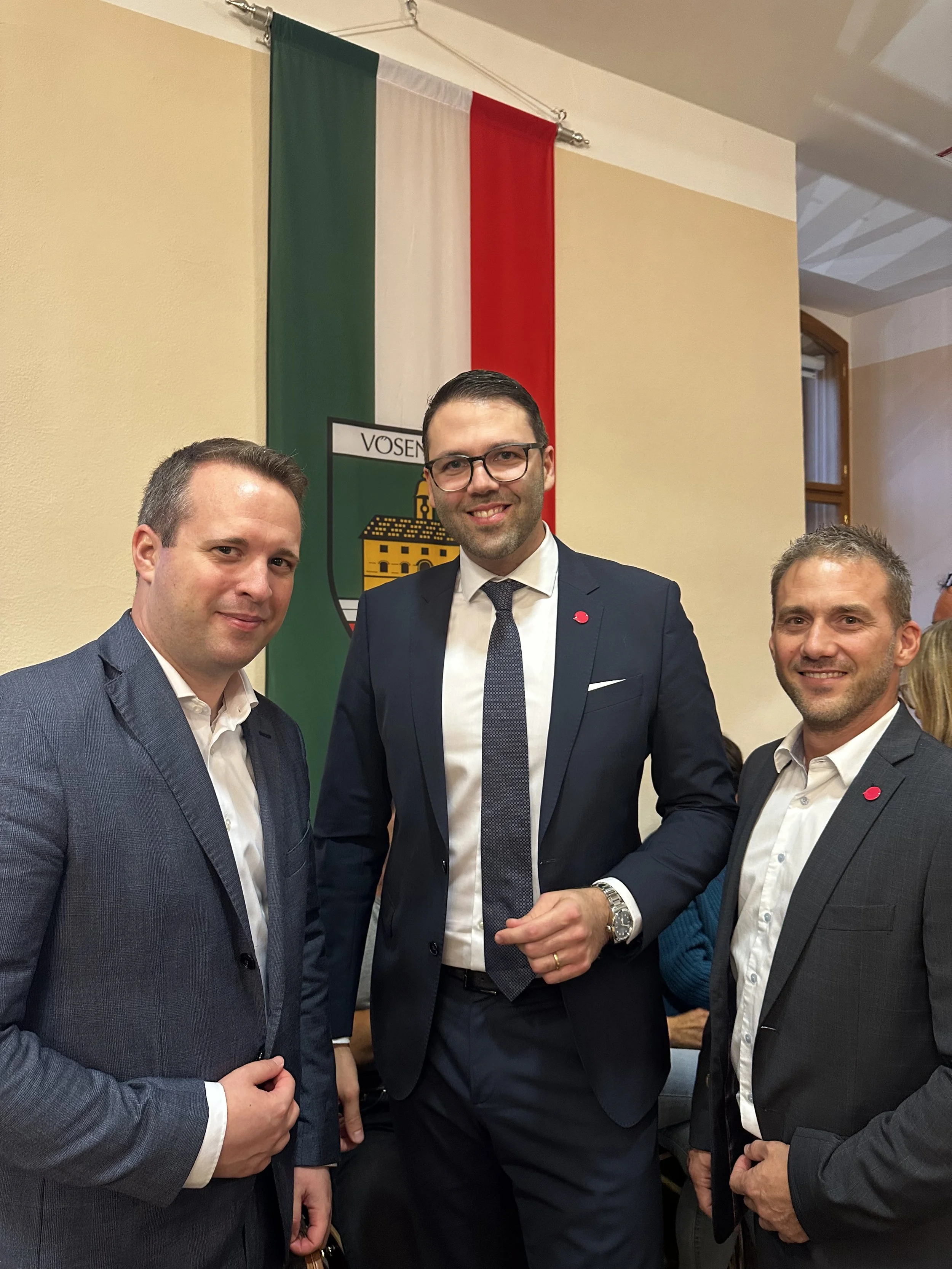 Drei Männer in Anzügen stehen vor einer Vösendorf-Flagge mit Wappen, die im Hintergrund hängt.