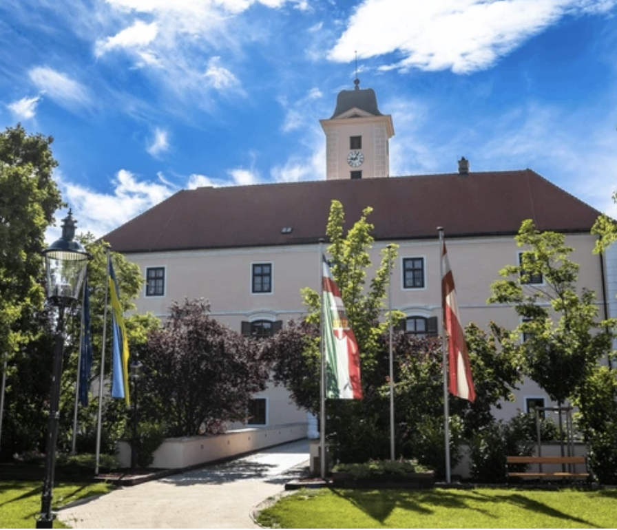 Schloss Vösendorf, umliegende Bäume, Flaggen auf Fahnenmasten, blauer Himmel mit einigen Wolken.