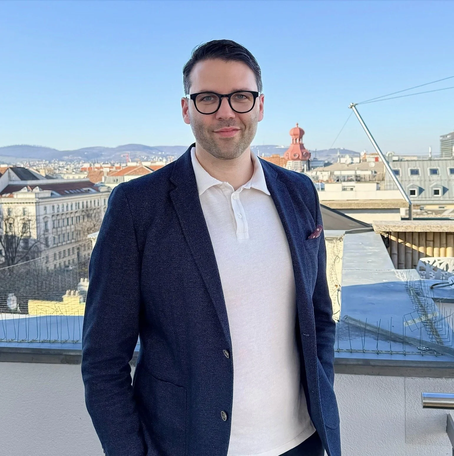 Ein Mann mit dunklen Haaren, Brille und einem weißen Poloshirt mit blauem Sakko steht auf einem Balkon mit Stadtblick, Himmel ist blau.