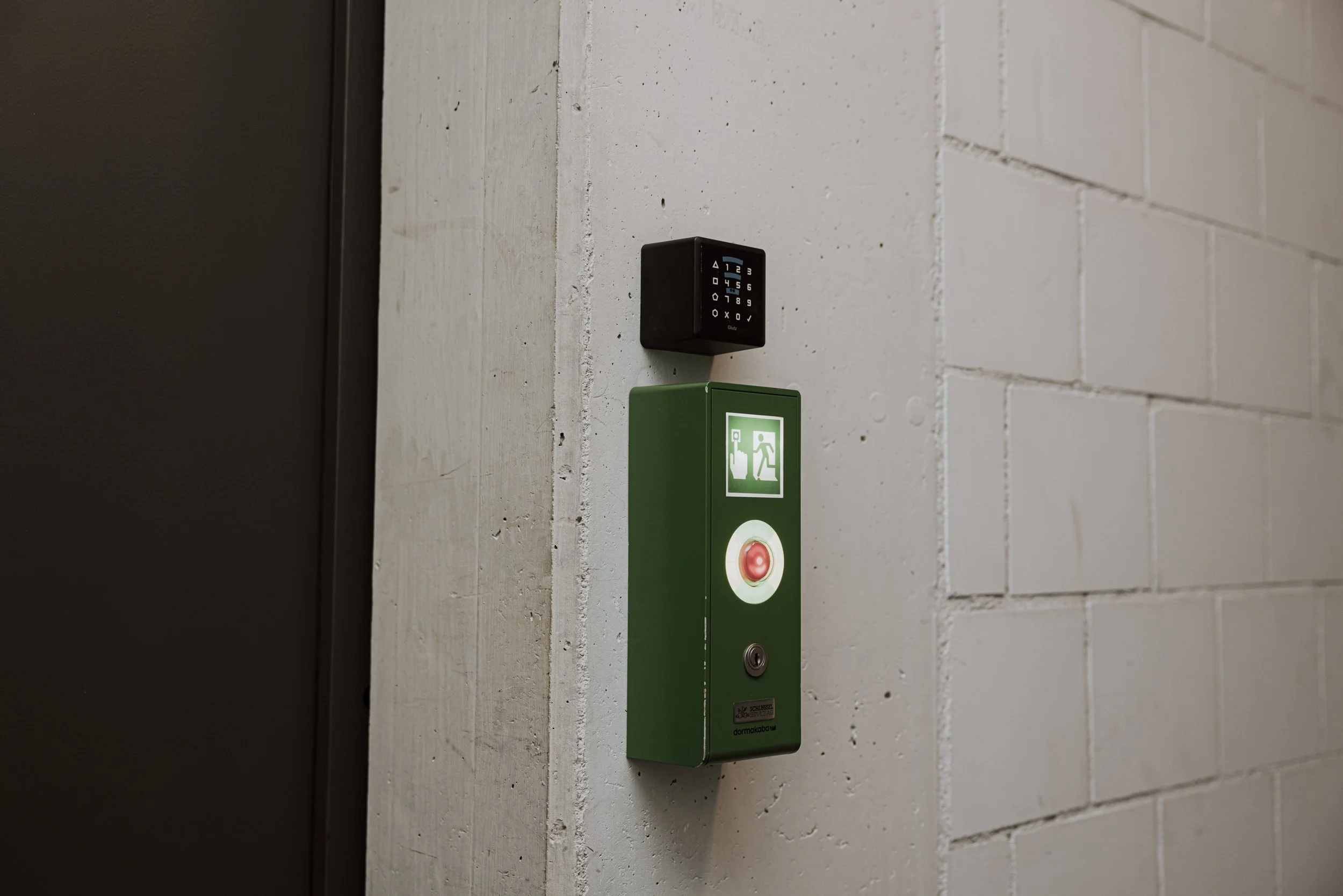 Notausgang mit grünem Notfallknopf und RFID-Zutrittskontrolle an einer weißen Ziegelmauer.