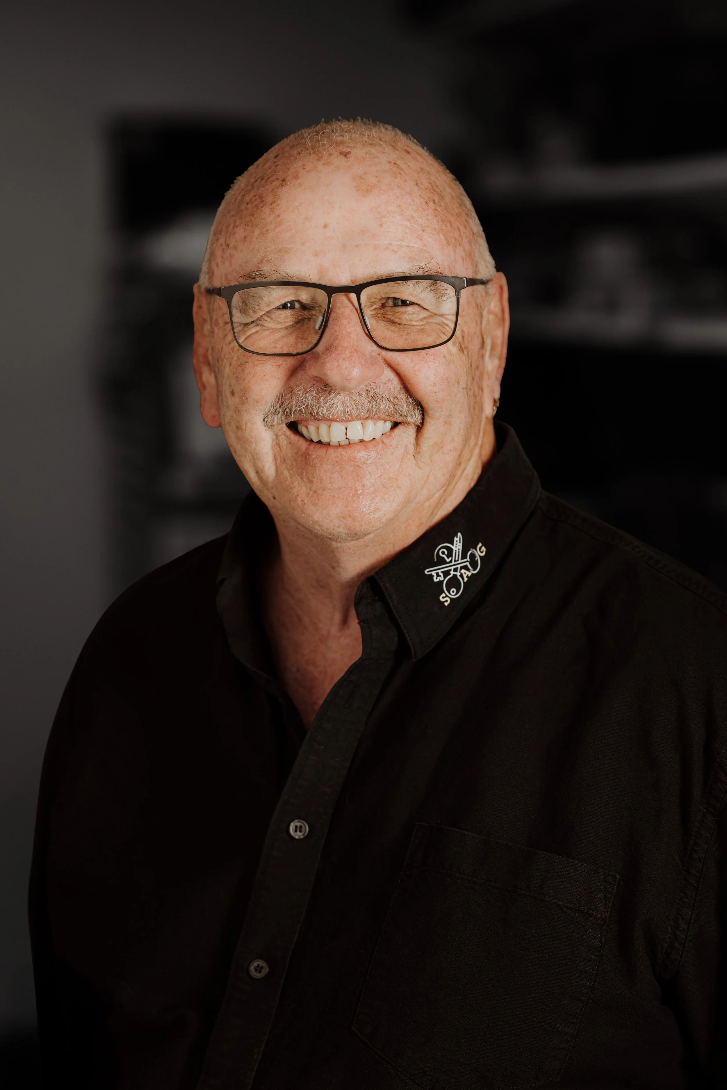 Lächelnder Mann mit Brille,grauem Haar, dunklem Hemd mit Logo auf der Kragen, im Hintergrund unscharf.