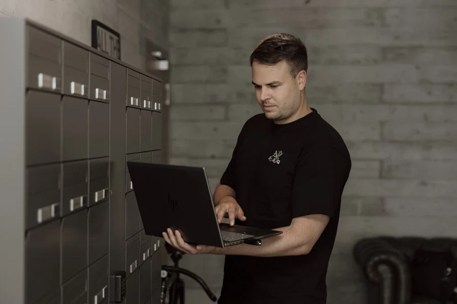 Ein Mann überprüft die Pakettauswahl in einem Postfachschrank, während er an einem Laptop arbeitet, in einem modernen Raum mit grauen Wänden.