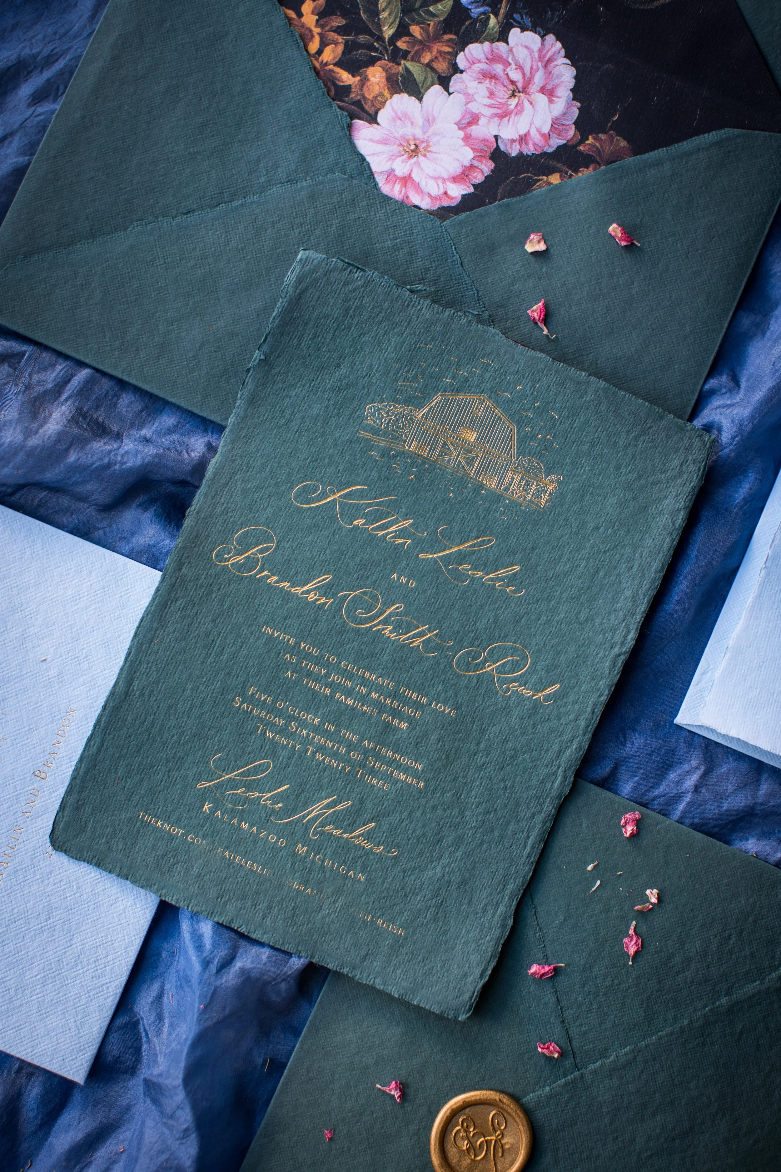 A wedding invitation card with ornate gold lettering and a small illustration of a barn, surrounded by matching dark green envelopes with pink flower petals and a gold wax seal, placed on a teal fabric background.