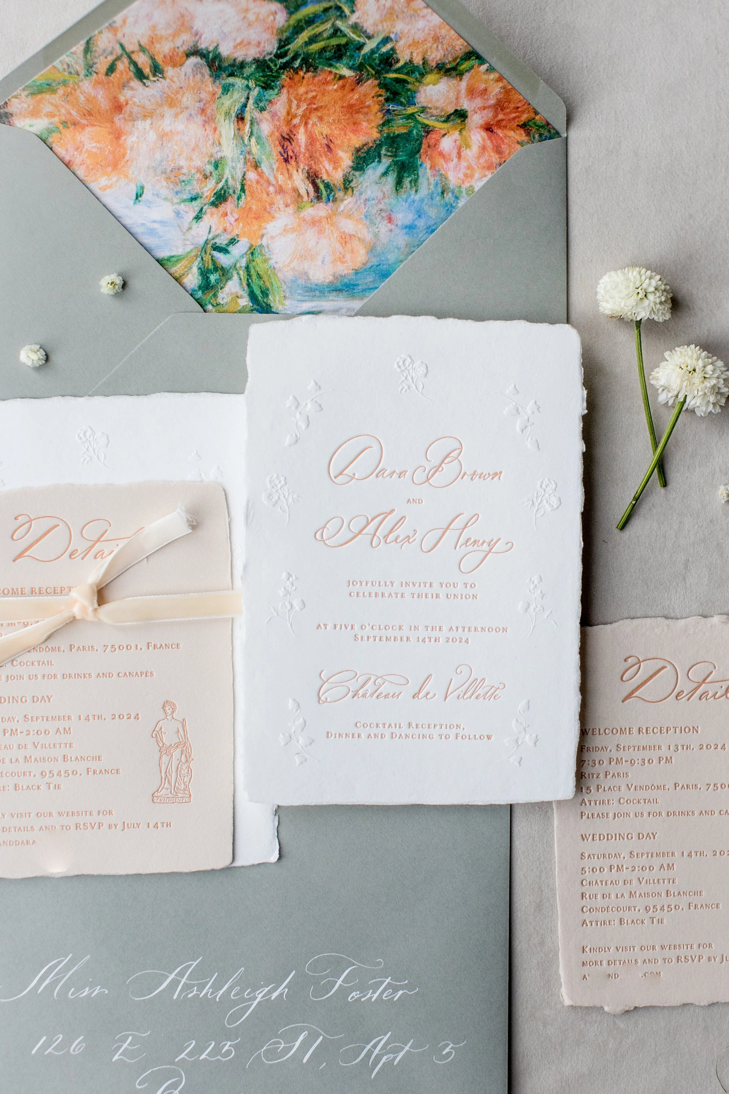 Wedding invitation suite with white paper embossed flowers, RSVP card with a cream ribbon, light gray envelope with floral liner, and small white flowers on a light gray background.