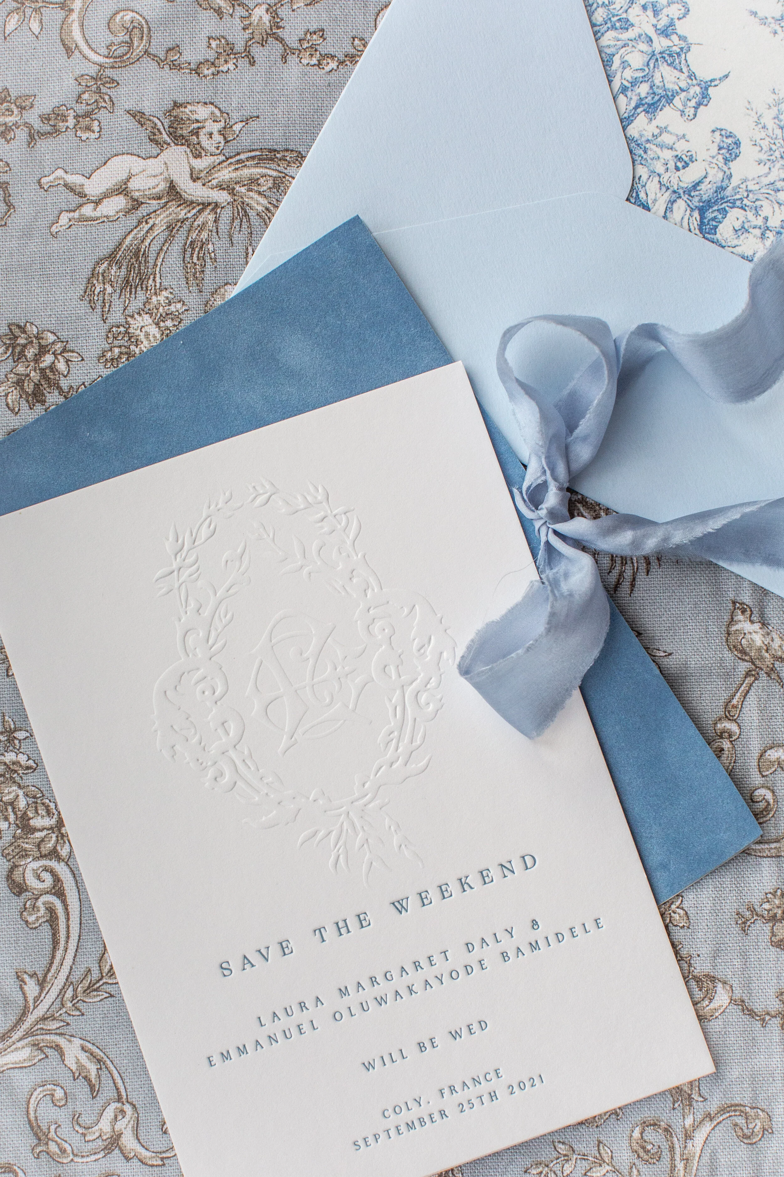A wedding invitation card with embossed monogram, placed on a blue envelope, tied with a satin ribbon, and set on a patterned tablecloth.