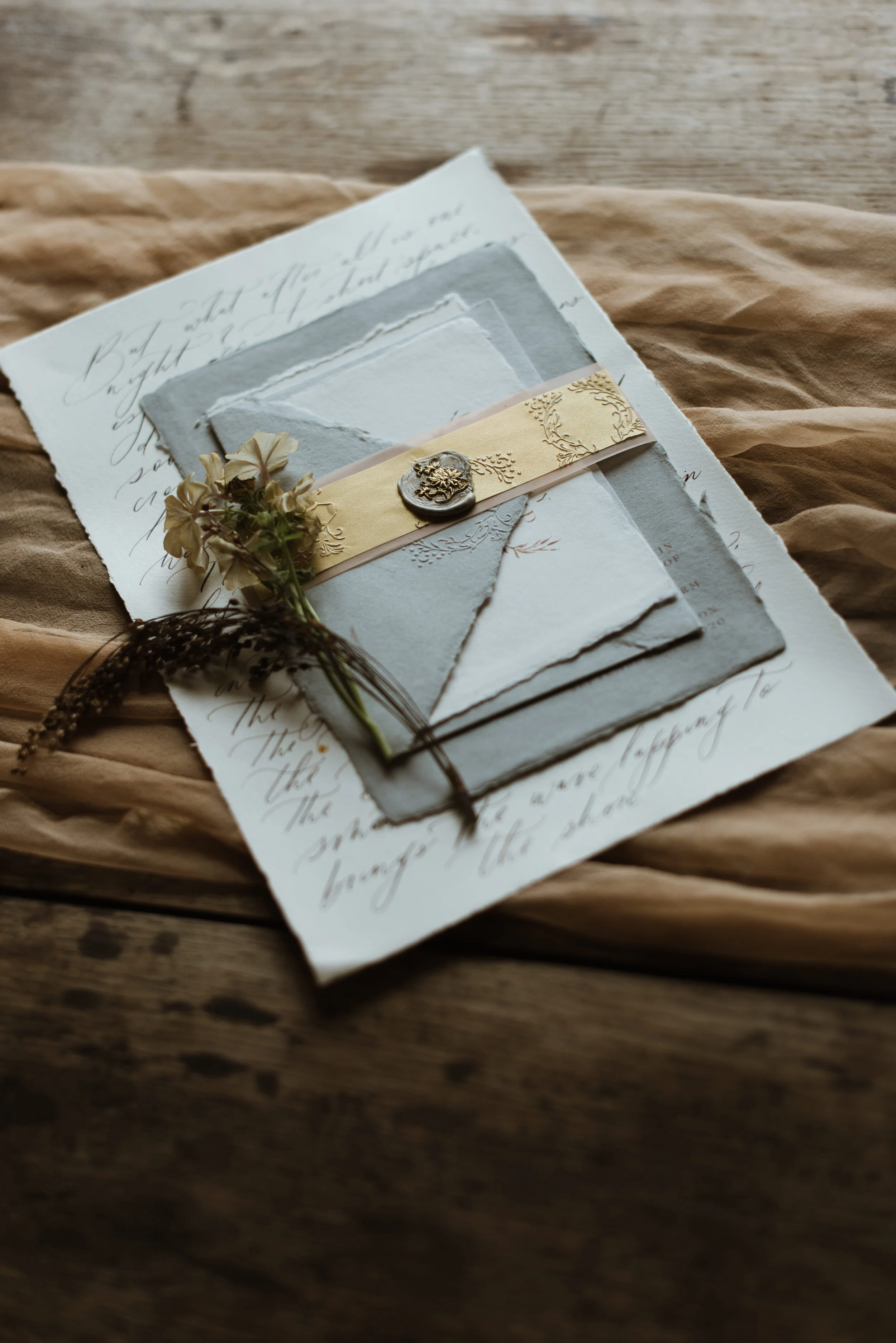 Vintage wedding invitation suite on rustic wooden table, including handwritten letter, textured paper, a paper band with decorative wax seal, and dried flowers.