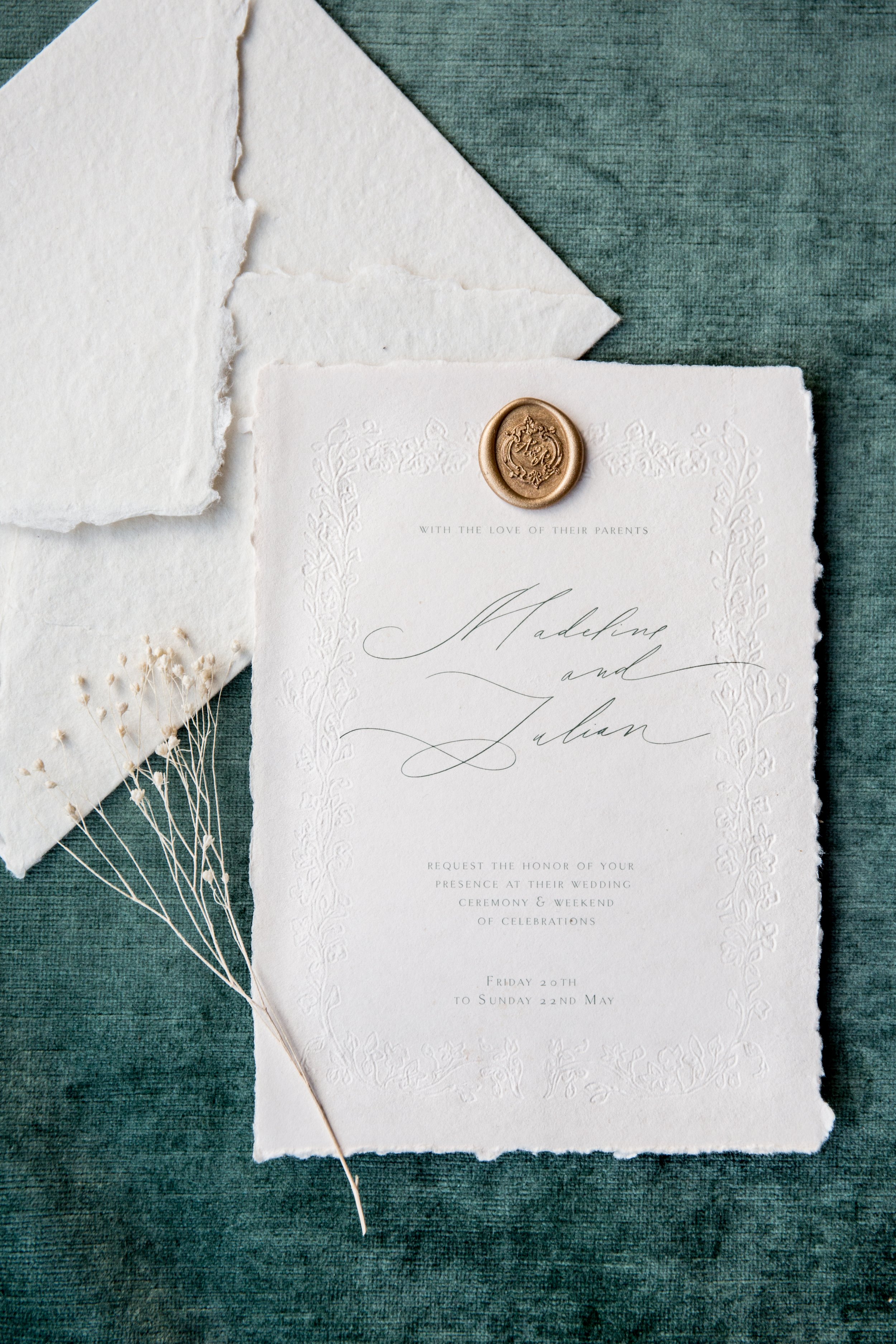 A wedding invitation with elegant script, a gold wax seal, and textured paper, set on a dark green fabric with additional torn paper and delicate dried flowers.
