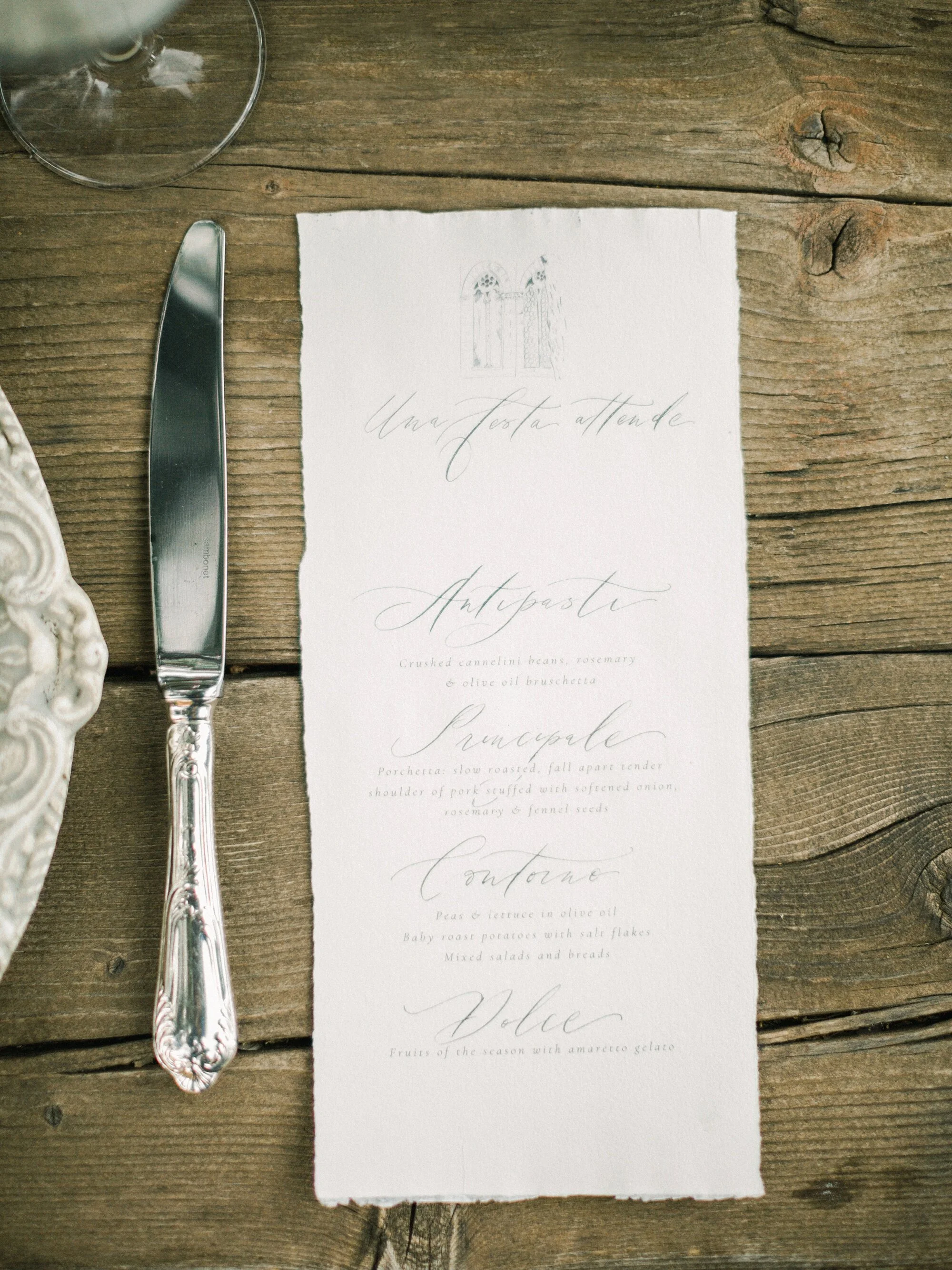 A handwritten style menu on textured white paper placed on a wooden table, with a silver-handled knife on the left and a glass of wine partially visible at the top.