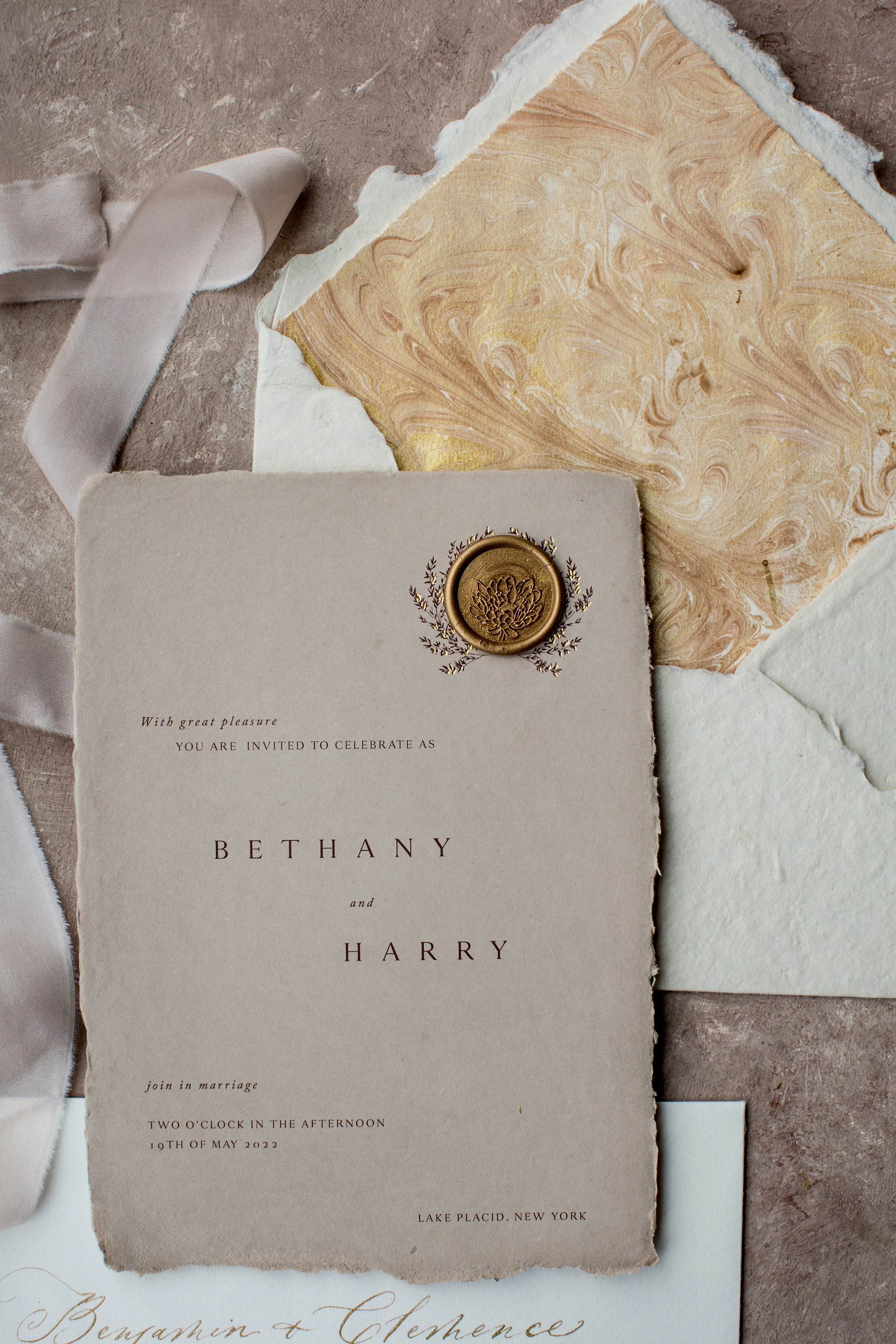 A wedding invitation with torn edges, featuring gold wax seal and floral design, placed on textured marble surface alongside textured paper, ribbon, and marble-patterned paper.