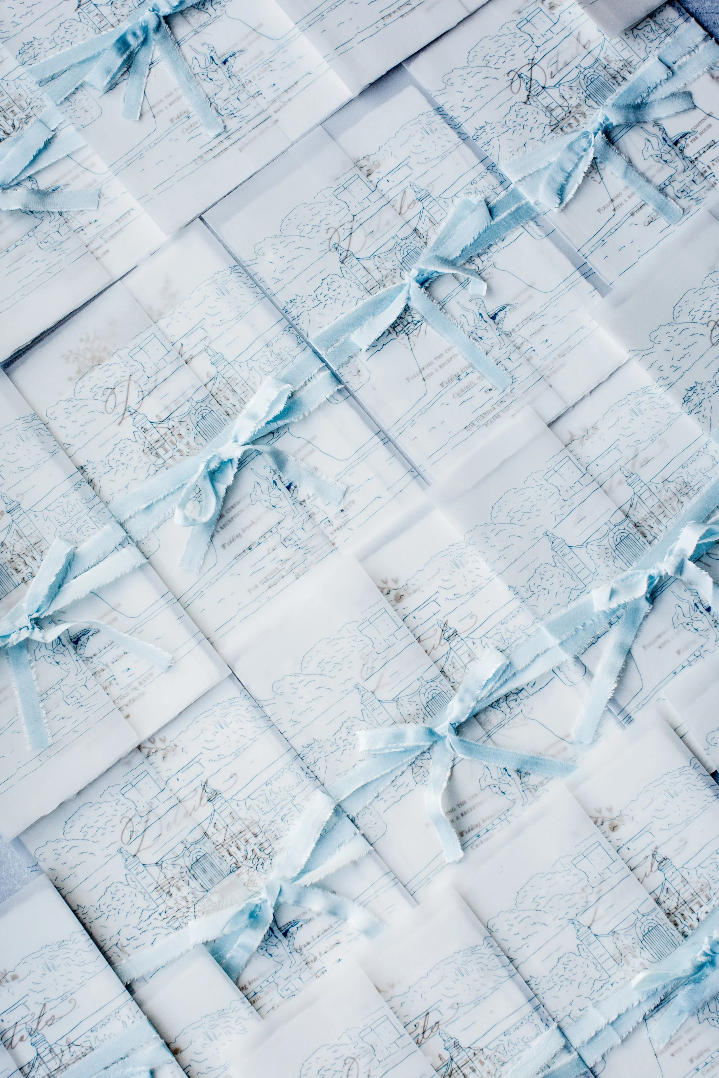 Multiple folded paper packages tied with satin ribbons, with delicate blue line drawings of landscapes or cityscapes, arranged in a grid pattern.