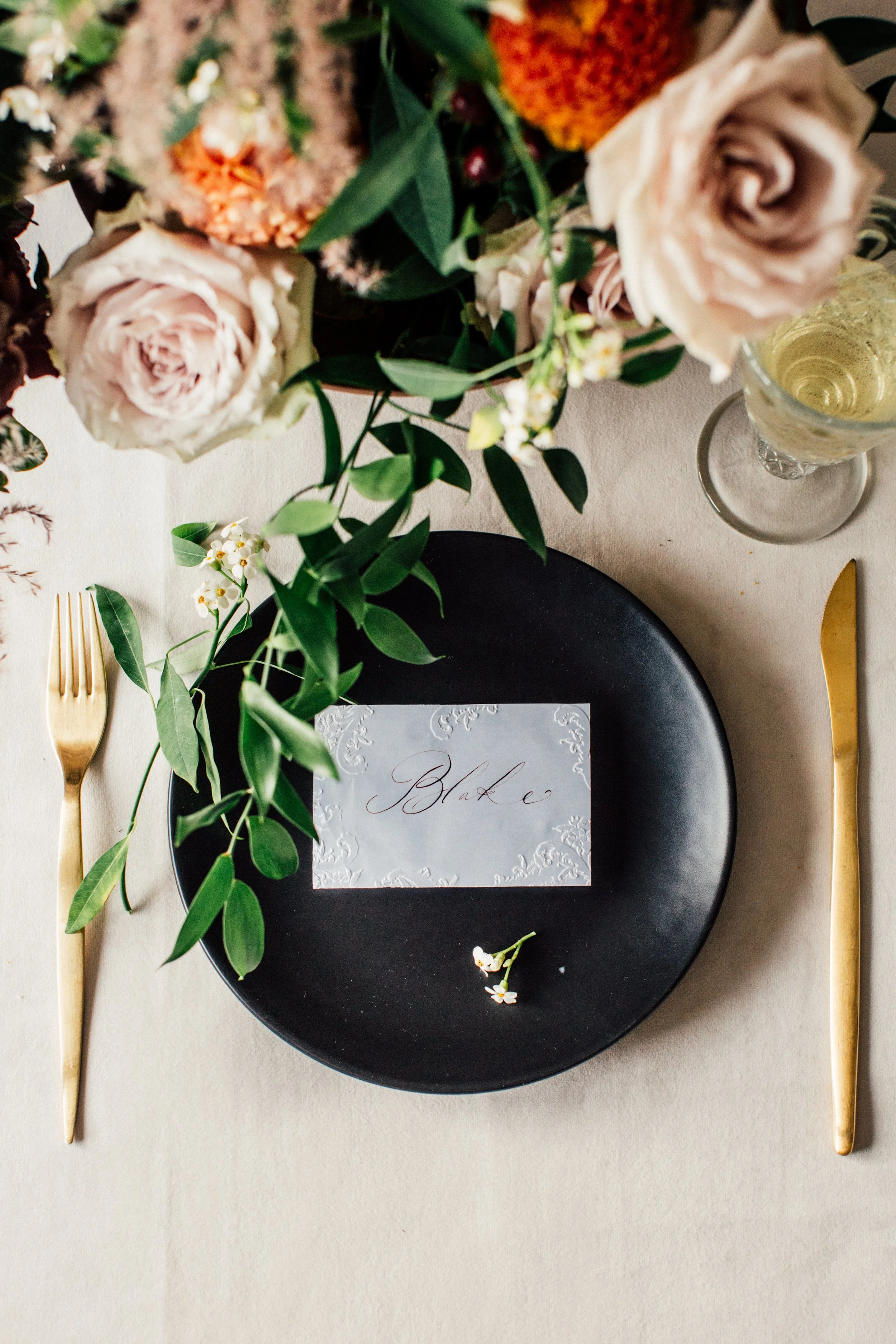 A table setting with a black plate, a white place card with the name 'Blake' written in cursive, gold utensils on each side, and a glass of white wine. A floral arrangement with roses, greenery, and other flowers decorates the table.