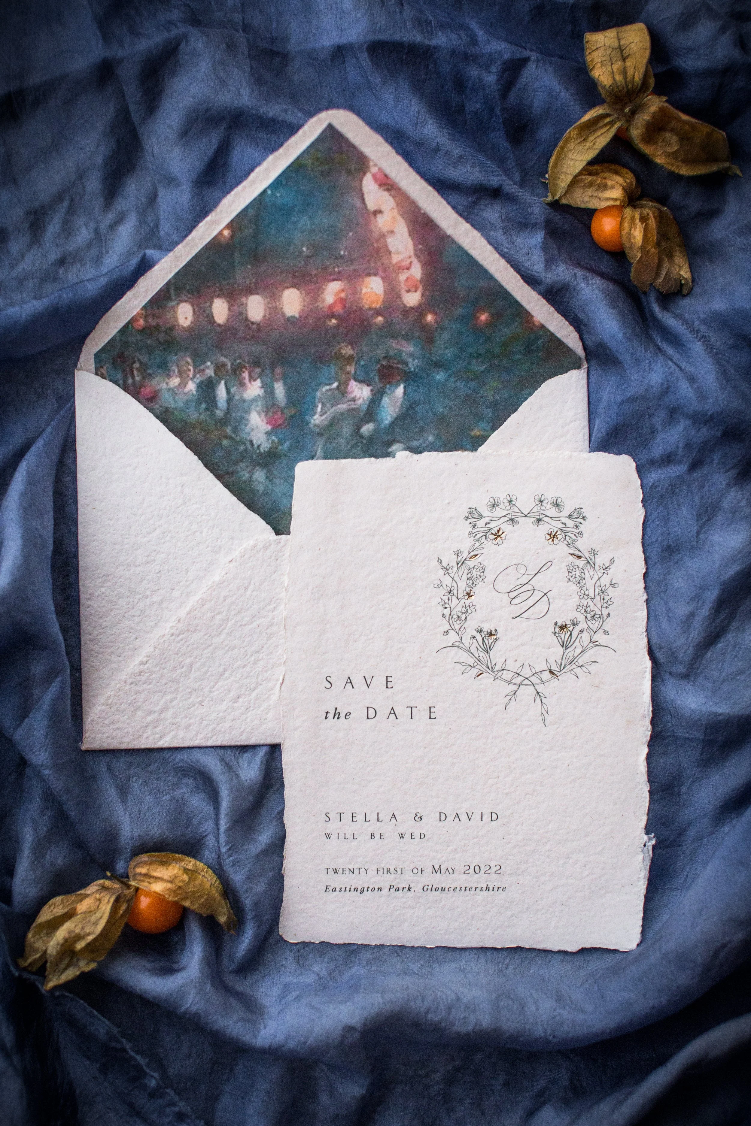 Wedding save the date invite with floral design and watercolor background, placed on navy fabric with dried physalis flowers nearby.