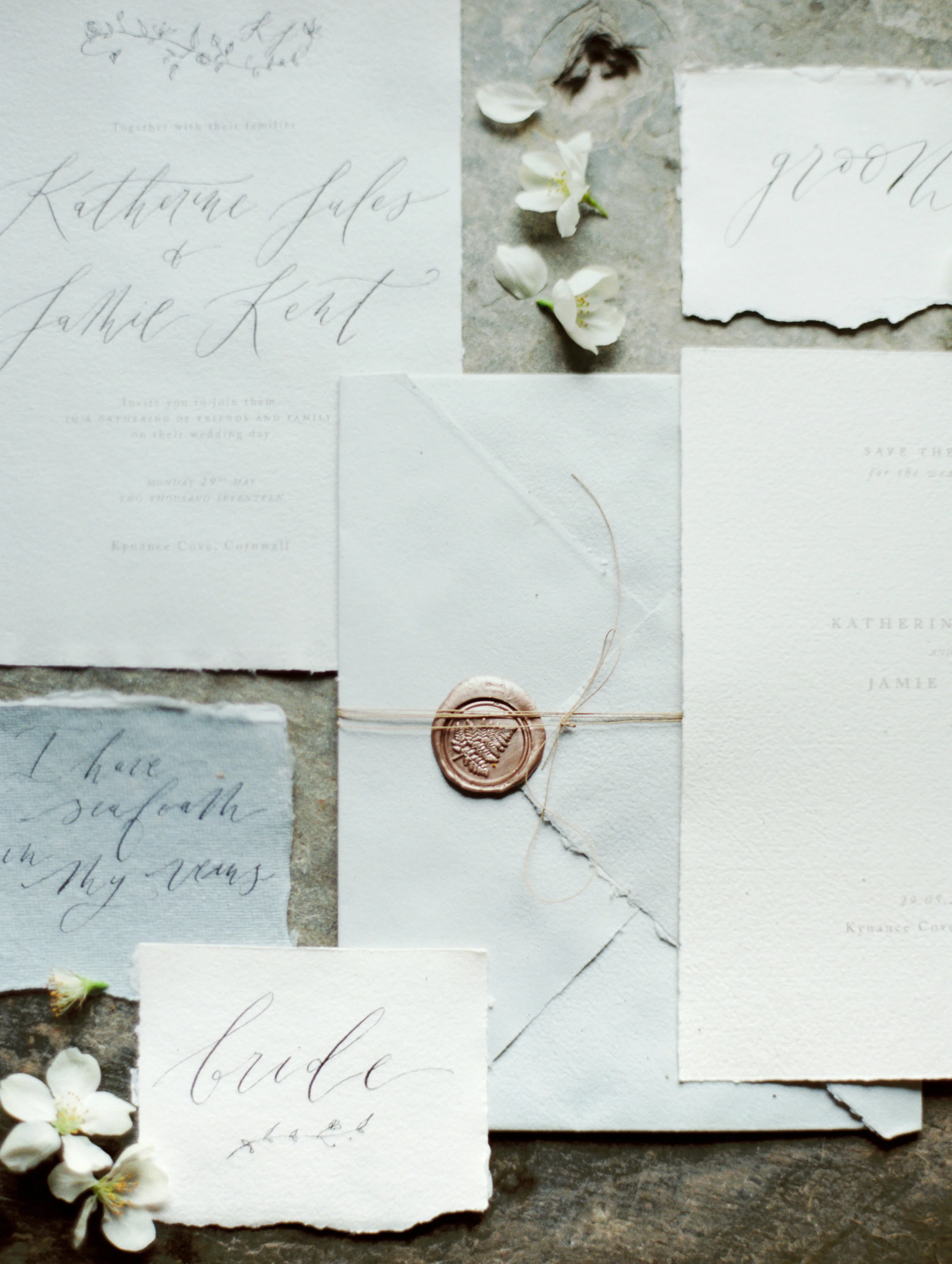 Wedding invitations, a sealed envelope with wax seal, and small white flowers arranged on a textured surface, all in neutral tones.