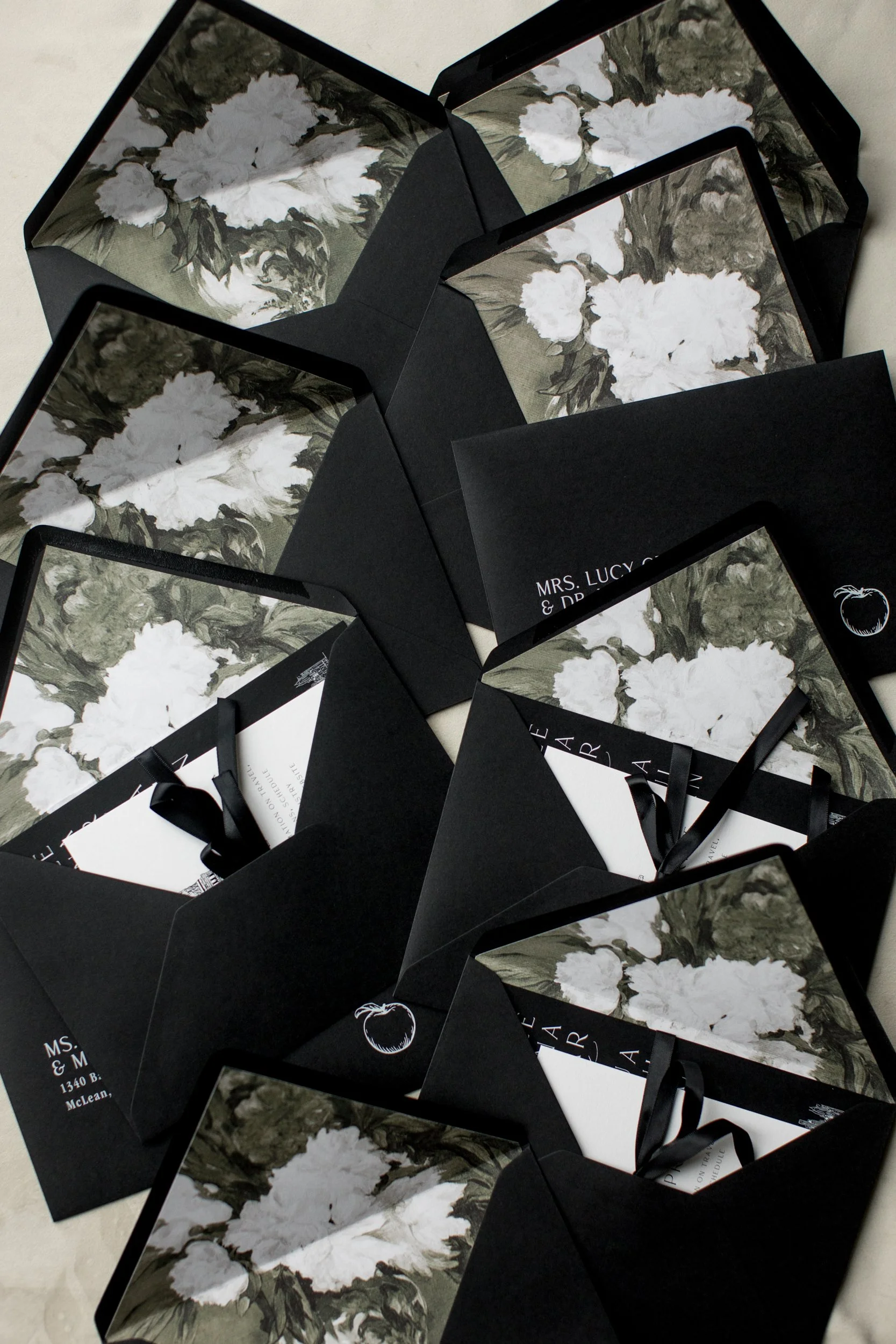 Several black invitation envelopes with floral liners featuring white flowers and green leaves, some with black satin ribbons, arranged on a flat surface.