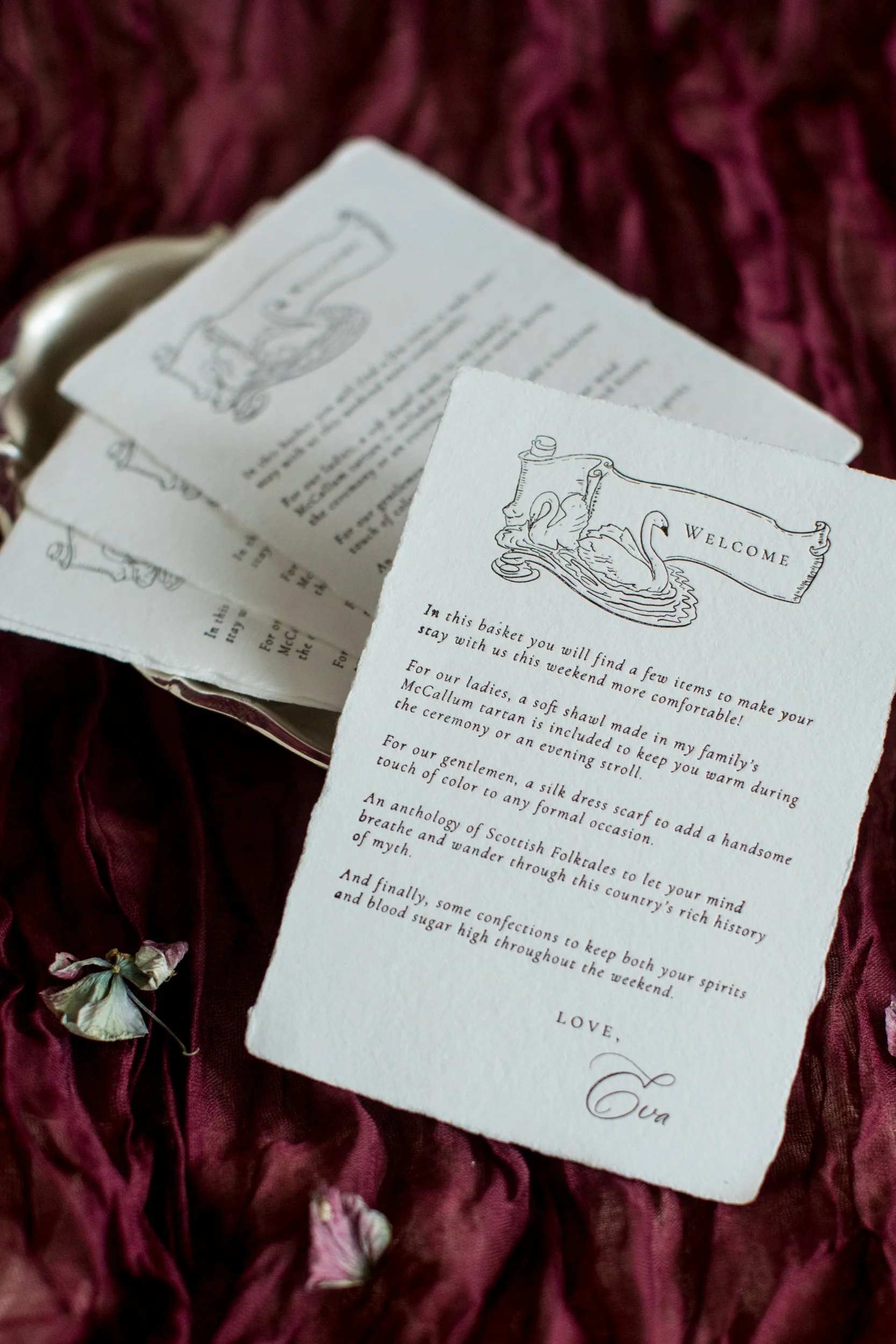 A set of vintage-style welcome cards with swan illustrations on a burgundy fabric background, accompanied by dried flowers.