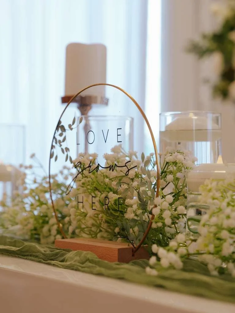 Decorative sign with the words 'Love grows here,' surrounded by white flowers and candles on a table.