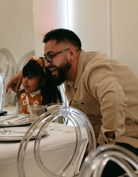 A man with glasses and a beard sitting next to a young girl with dark hair and a bow, smiling at a dining table.