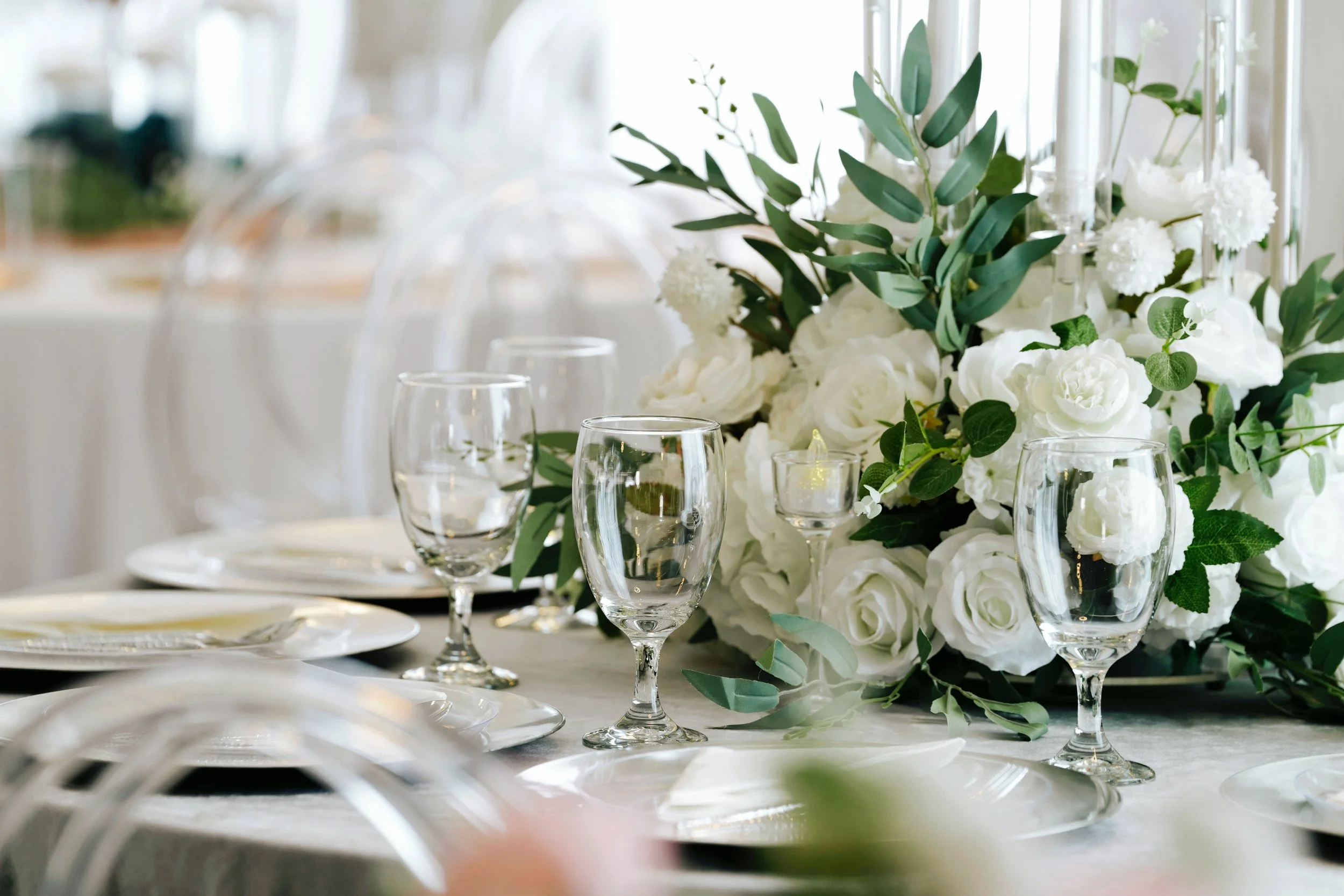 Elegant table setting with a floral centerpiece of white roses, green leaves, and candles, surrounded by glassware and plates for a formal event.