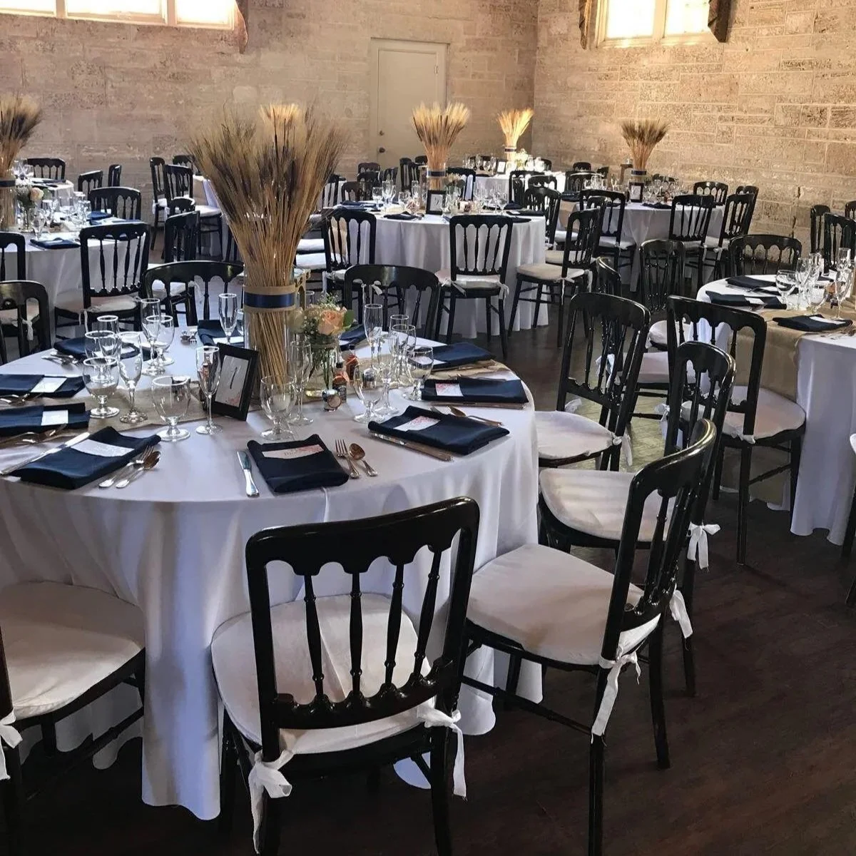 Round tables set with white tablecloths, black and white chairs, elegant glassware, and black napkins, decorated with tall dried wheat centerpieces in a rustic brick-walled venue, prepared for a formal event.