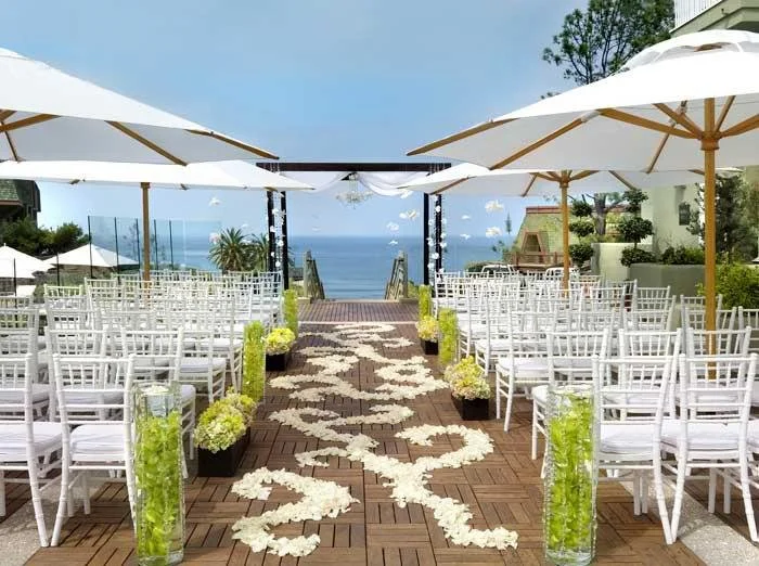 Outdoor wedding ceremony setup with white chairs, floral aisle, umbrellas, and ocean view.