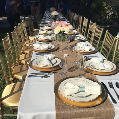 Long rectangular outdoor banquet table set with white tablecloth, gold chairs, floral centerpiece, and place settings including plates, glasses, napkins, and cutlery.