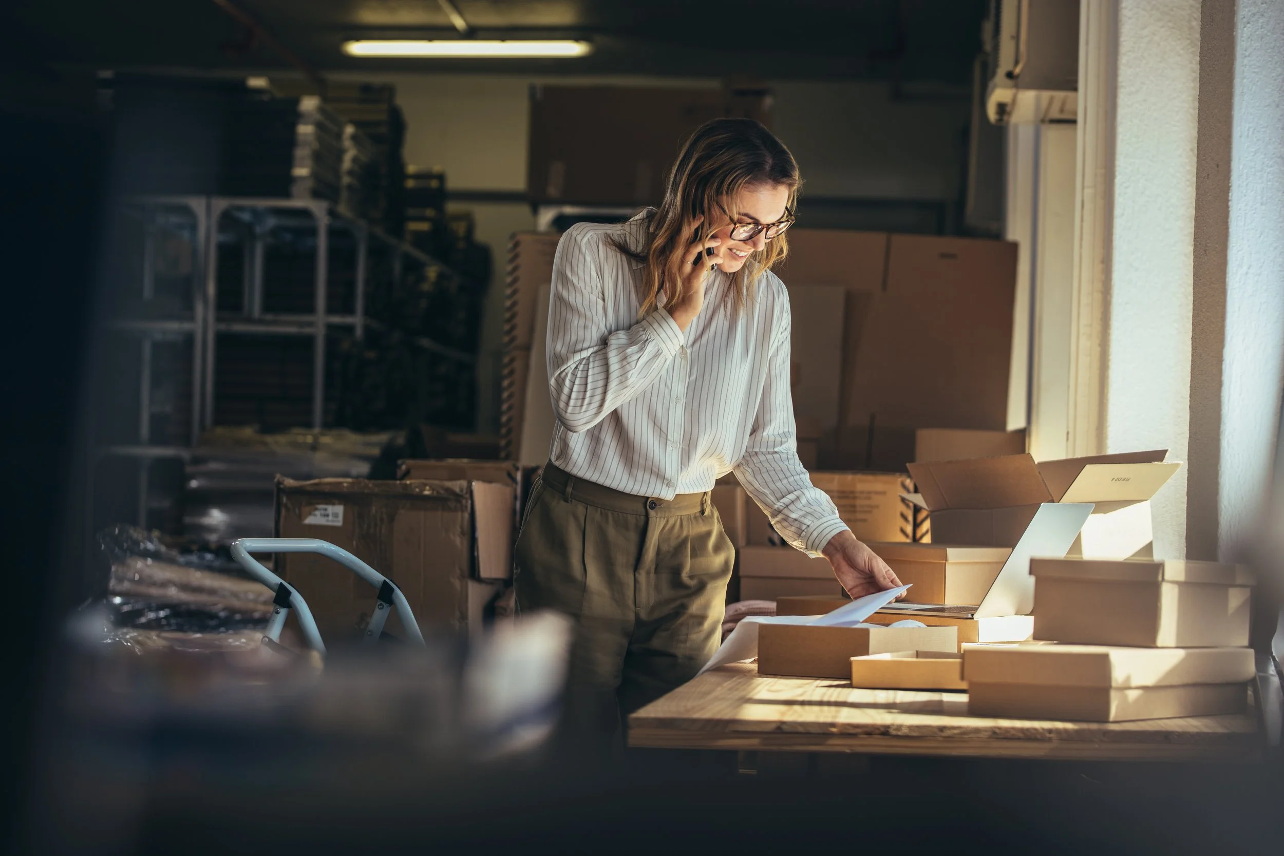 A woman wearing glasses and a striped blouse smiles while talking on her cell phone and looking at documents on a desk covered with cardboard boxes in a warehouse or storage room.