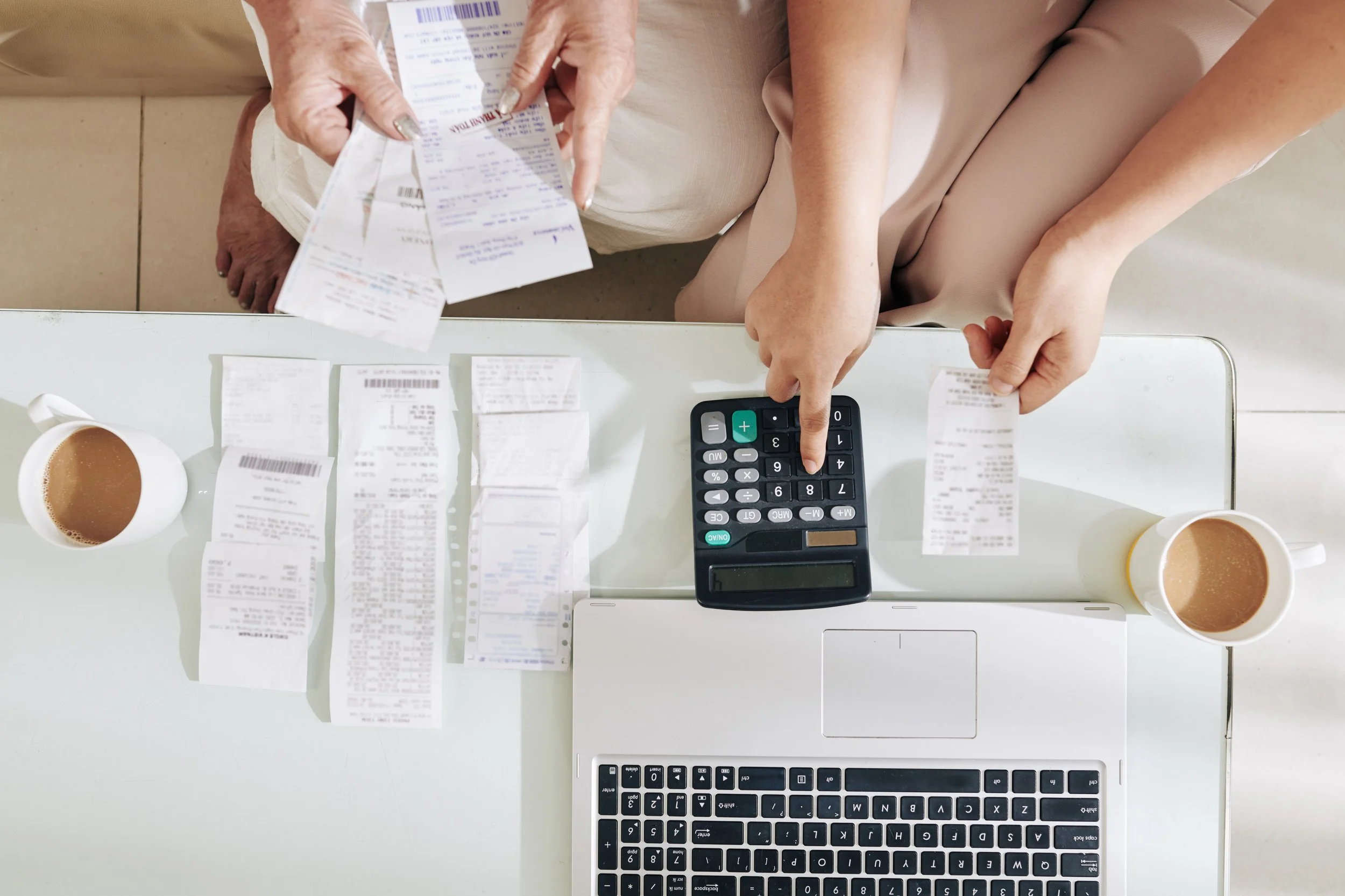 Two people are sitting at a white table with a laptop, a calculator, and several receipts spread out. Each person is holding a receipt, and there are two cups of coffee on the table.