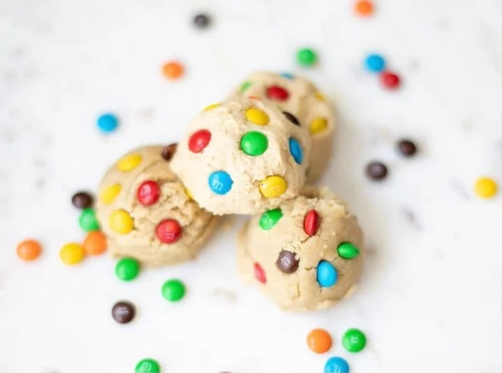 Chocolate chip cookie with colorful candy-coated chocolates on top, surrounded by scattered similar candies on a white surface.
