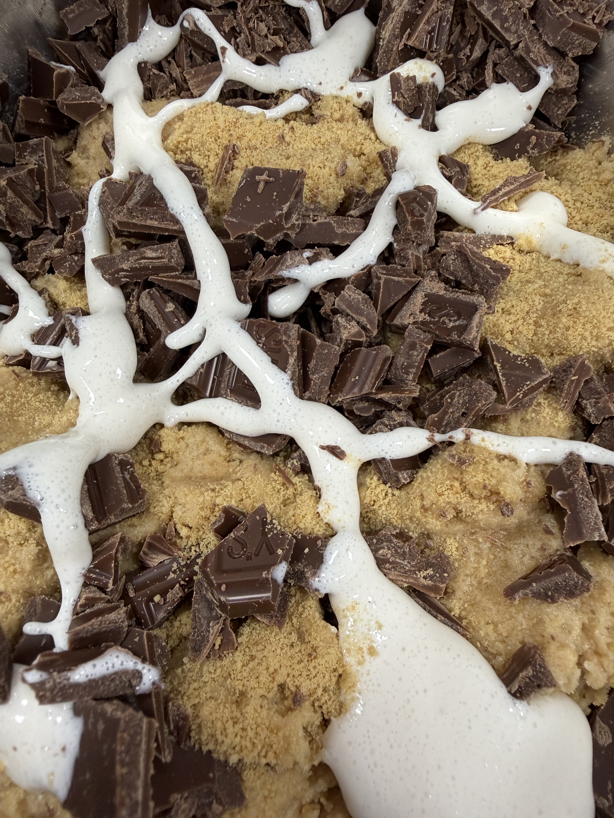 Close-up of a dessert with broken chocolate pieces, light-colored crumbs, and white sauce or cream drizzled on top.