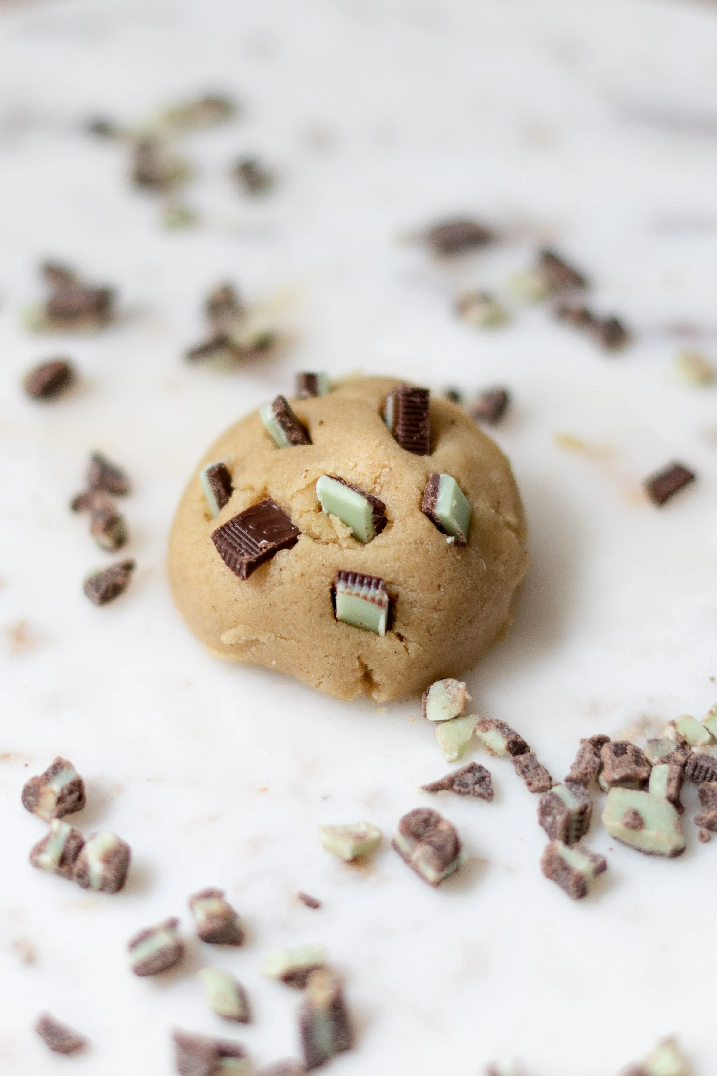 Chocolate chip cookie dough ball with chopped chocolate and mint candies scattered around on a white surface.