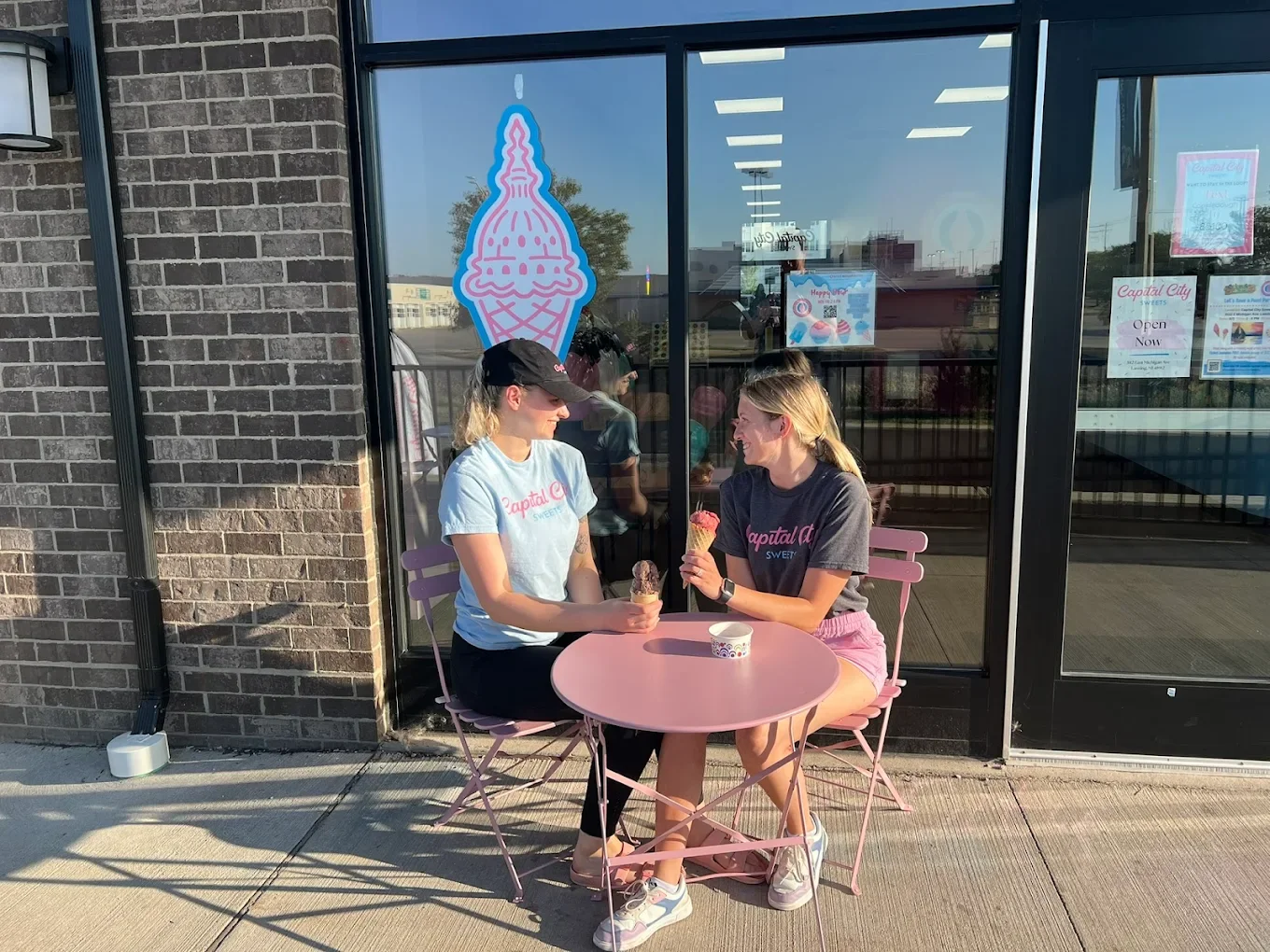 Two women sitting outside an ice cream shop, sharing a conversation and holding ice cream cones. Behind them is a large pink ice cream cone logo on the window of the shop.