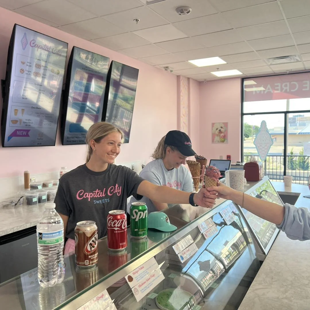 Two women working behind the counter at a soft serve ice cream parlor, handing an ice cream cone to a customer. There are ice cream flavors, soda cans, and a water bottle on the counter.