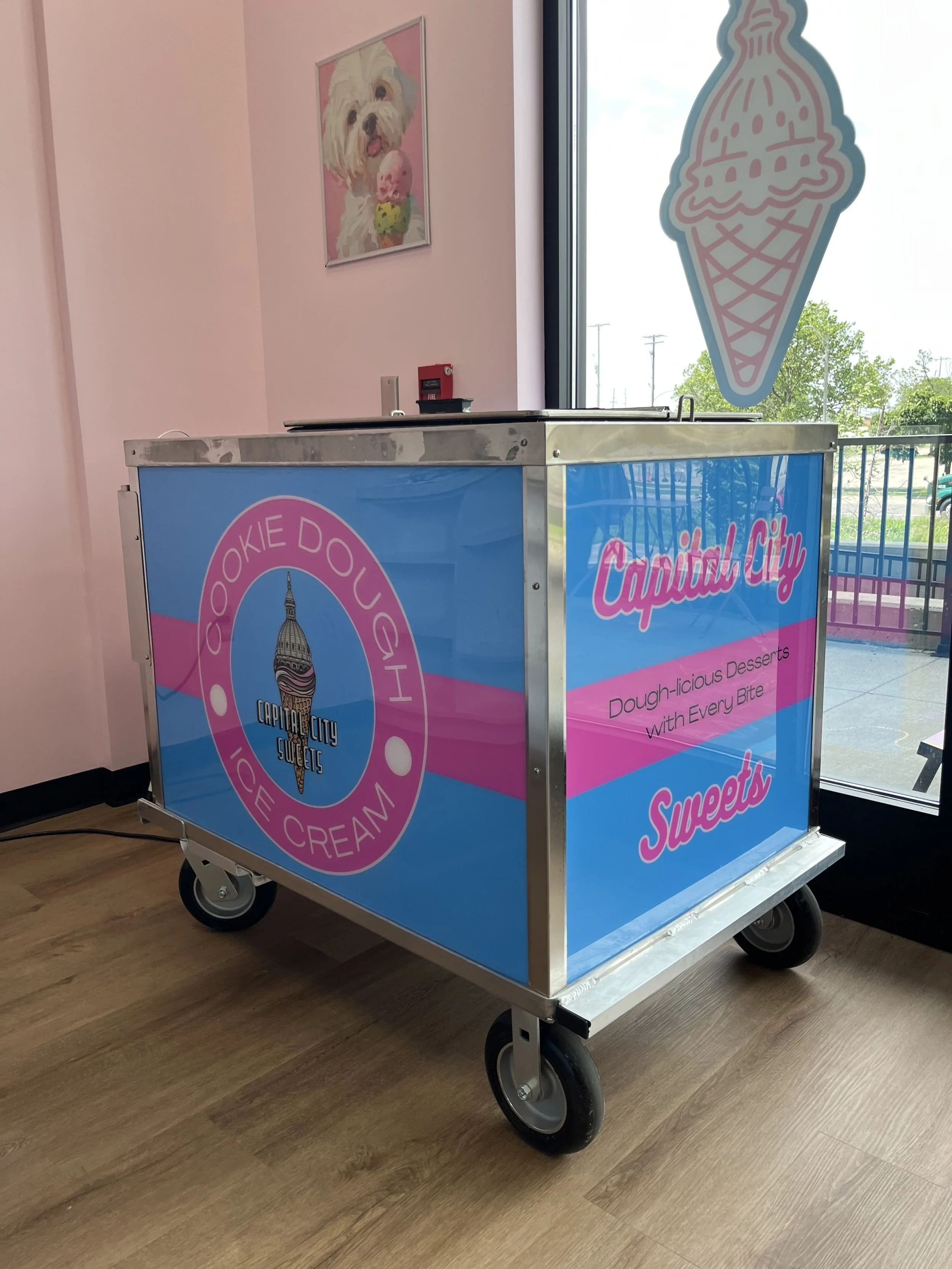 Ice cream cart with branding for Cookie Dough Sweets in a pink and blue color scheme, located indoors near a glass door, with a pink wall and dog portrait hanging on it.
