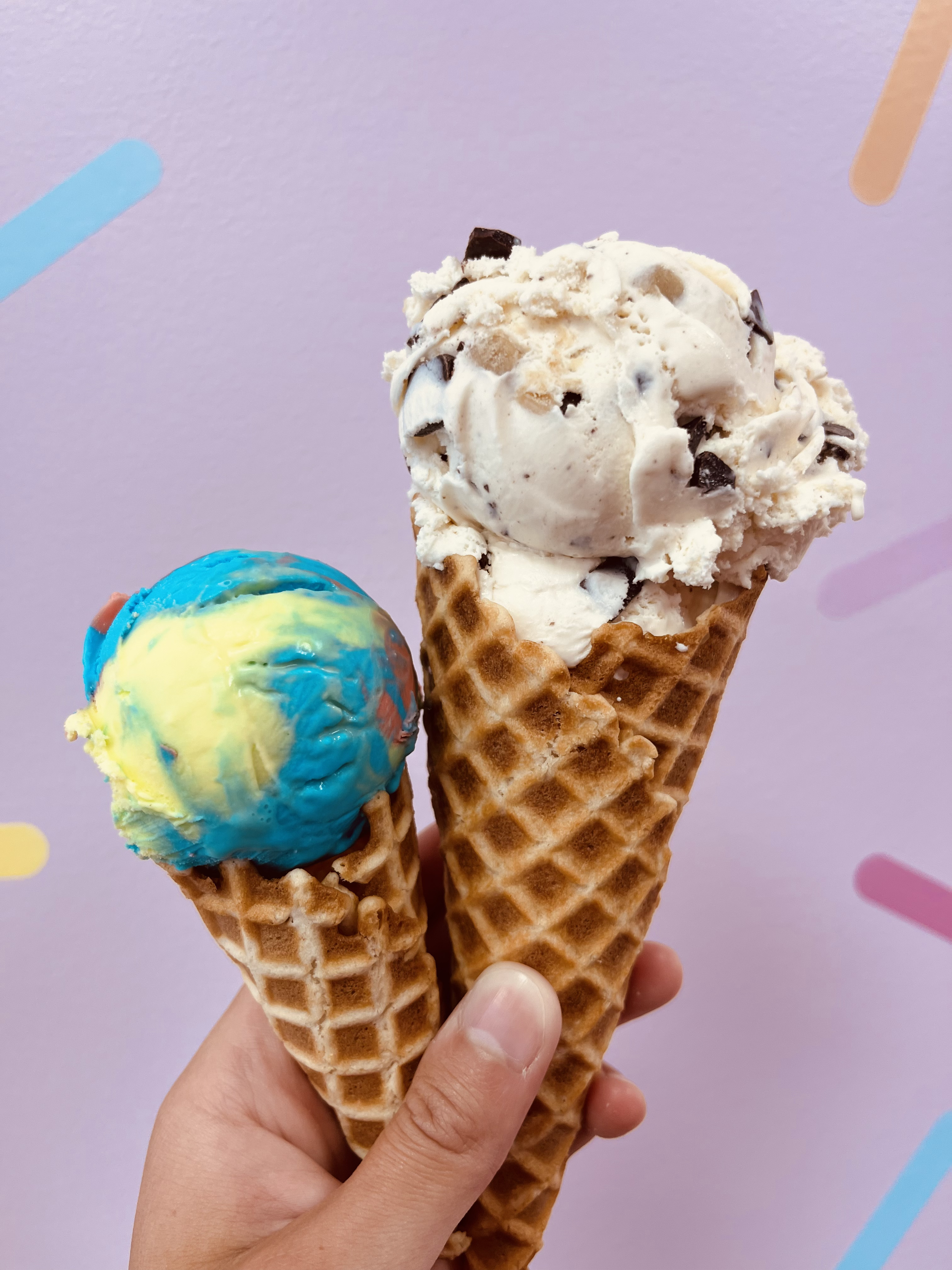 A hand holding two waffle cones with ice cream, one chocolate chip and vanilla in a waffle cone and the other colorful blue, yellow, and red in a waffle cone, against a pastel purple background with colorful sprinkles.