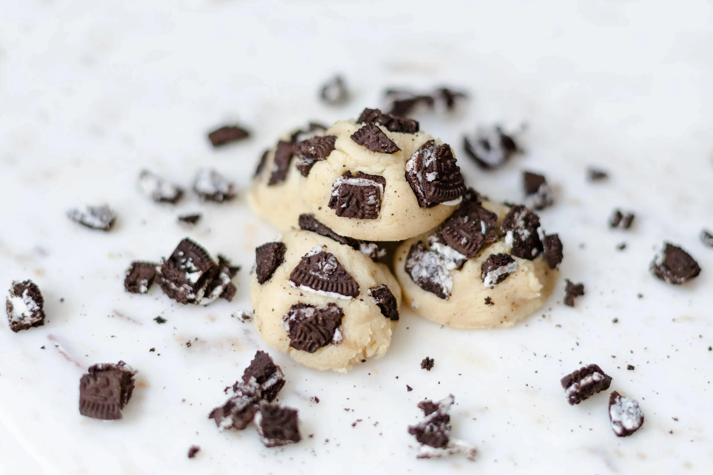A close-up of a cookie with chocolate and green candy pieces on a white surface with scattered chopped chocolates.