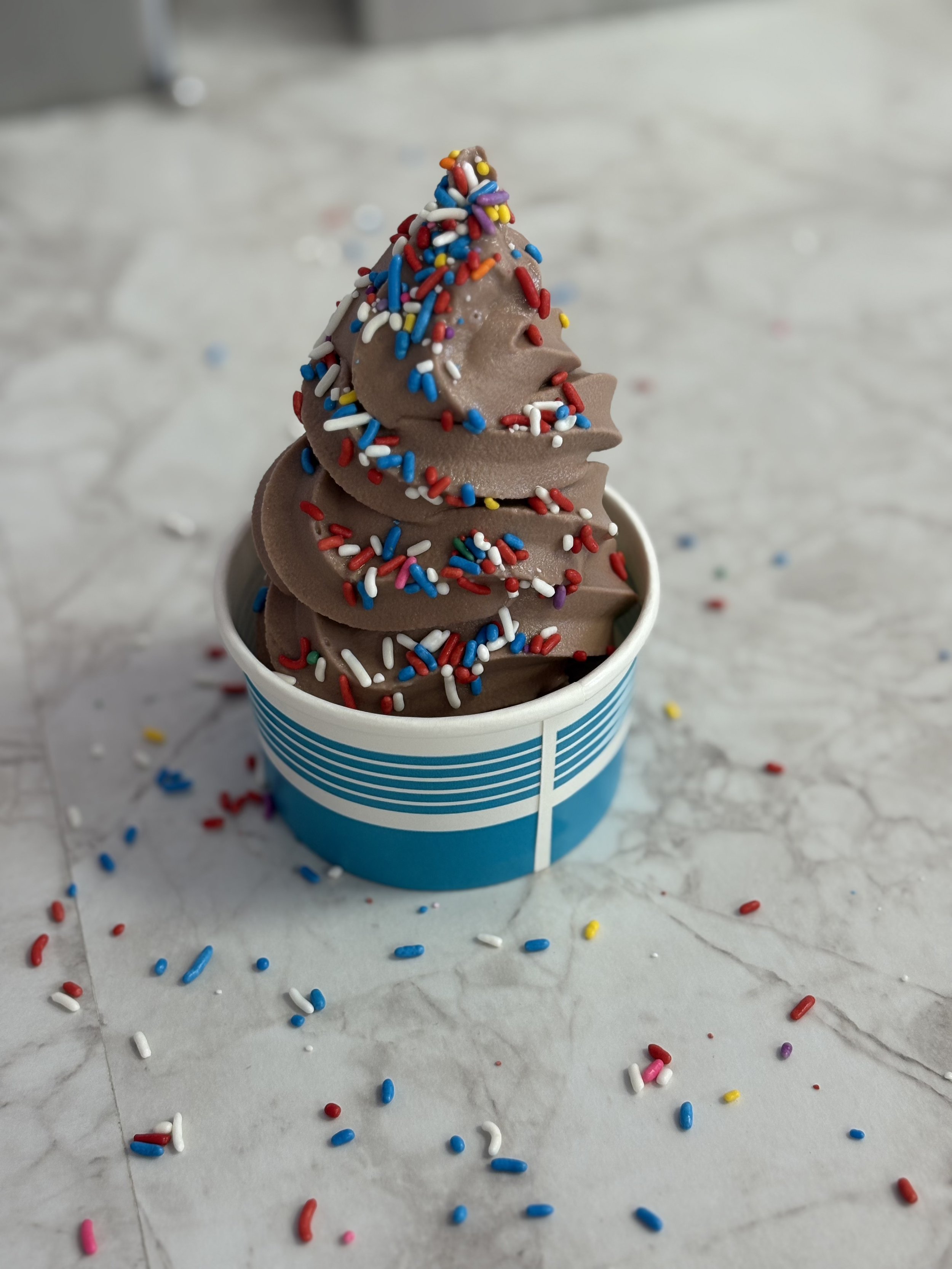 Chocolate soft serve ice cream with rainbow sprinkles in a blue and white striped cup on a marble surface