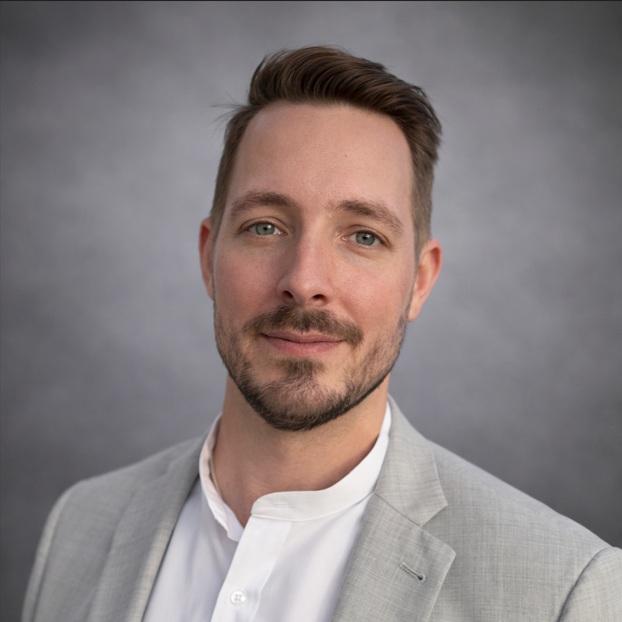 A man with light skin, short brown hair, and a beard, dressed in a light grey blazer and white shirt, smiling against a grey background.