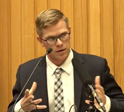 A man in a suit with glasses speaking at a microphone, in front of a wooden panel background.