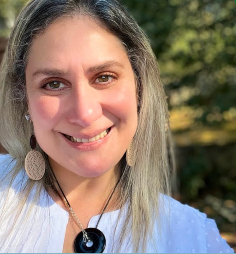 A woman with blonde hair and brown eyes smiling outdoors. She is wearing large beige earrings and a black necklace with a circular pendant, along with a white top. The background shows green foliage.