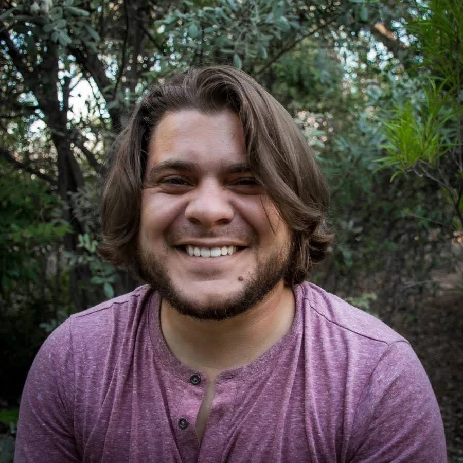 A smiling man with shoulder-length brown hair and a beard, wearing a purple shirt, outdoors in a wooded area.