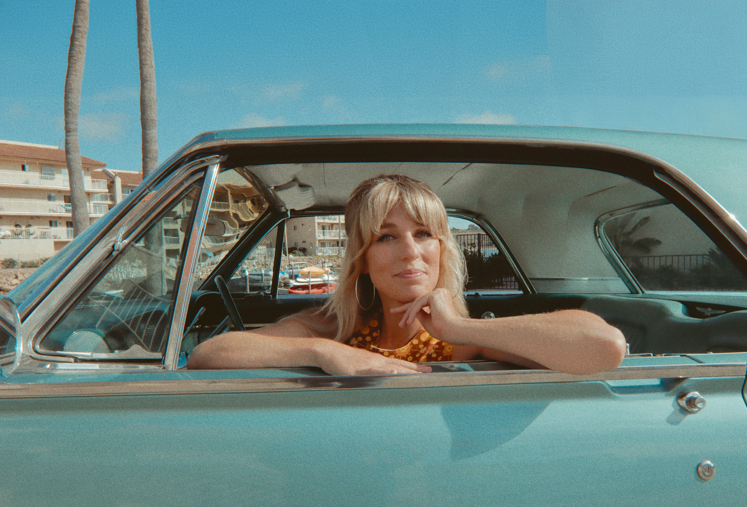 Country singer Kimmi Bitter sitting in a vintage turquoise car, resting her chin on her hand, with a marina and buildings in the background under a blue sky.