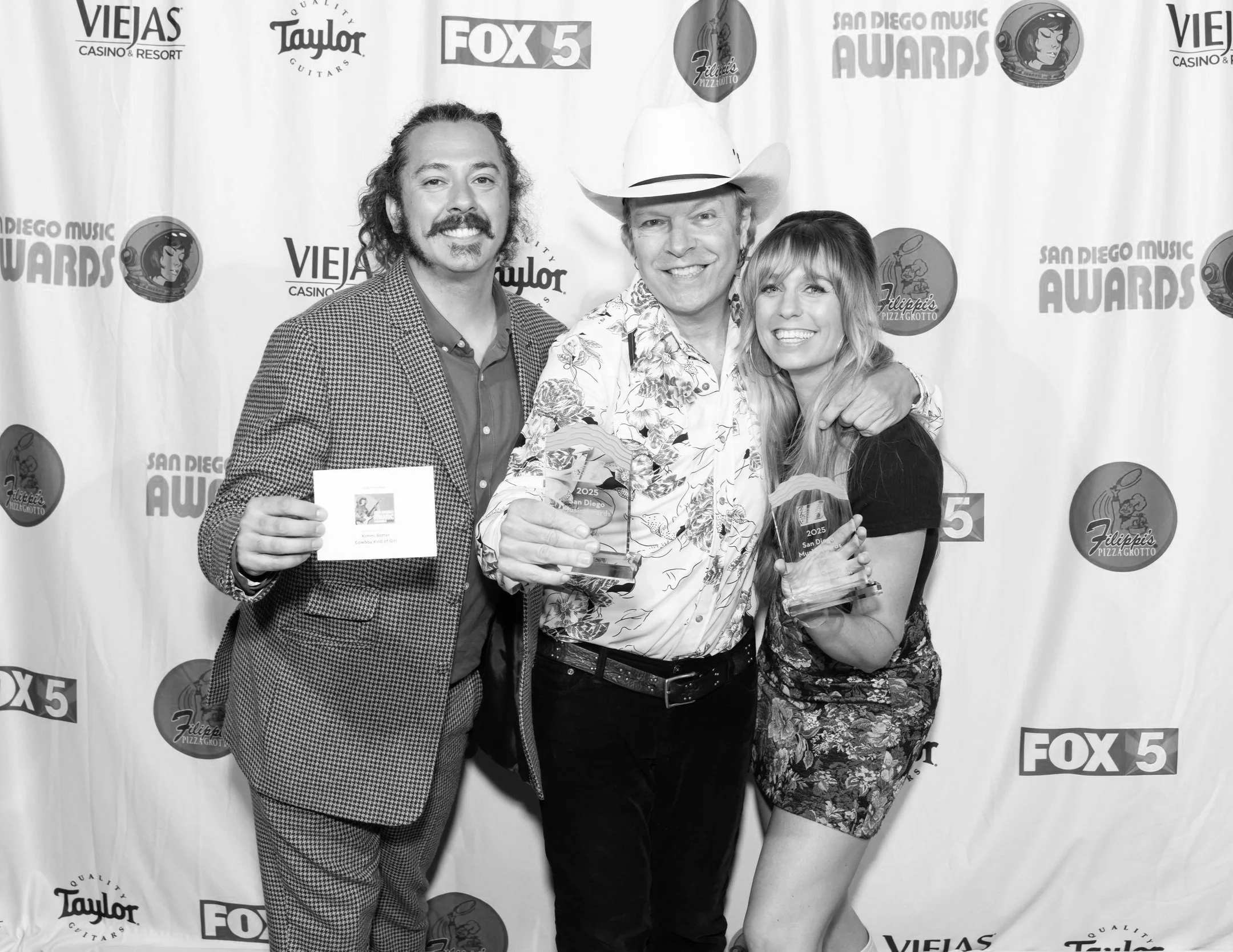 Kimmi Bitter and crew standing together at an award event, smiling, with a backdrop featuring various sponsor logos for 'San Diego Music Awards'. The person in the middle is holding an award, and the person on the right is also holding a trophy.