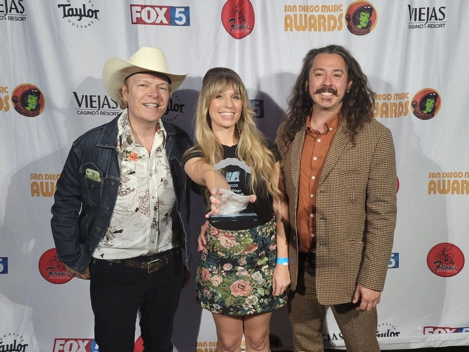 Kimmi Bitter, Michael Gurley, And Willis Farnsworth smiling at an awards event, holding trophies and a certificate, standing in front of a backdrop with logos including FOX 5, Viejas, and San Diego Music Awards.