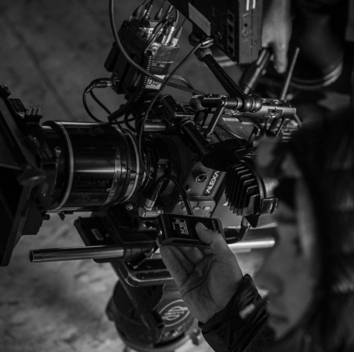 Black and white photo of a professional film camera mounted on a tripod, with a person adjusting settings using a smartphone.