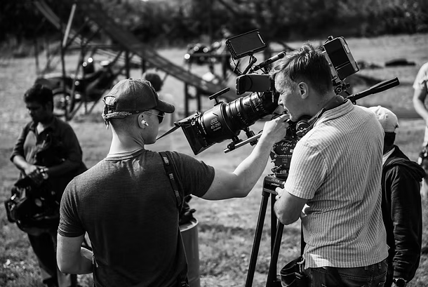 Two men operating a professional camera on a film or video set, with others in the background and outdoor equipment visible.